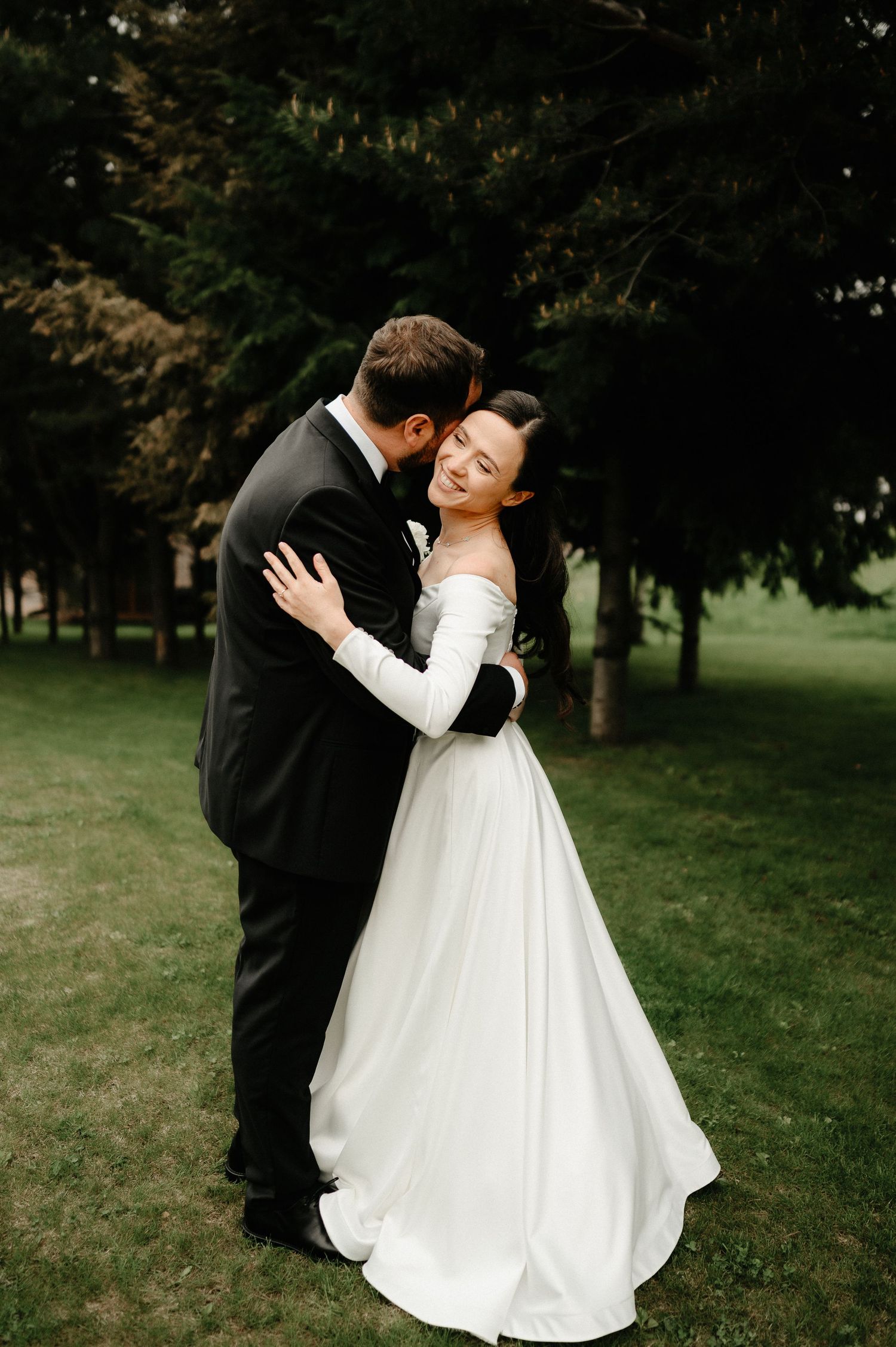 Smiling bride in off-shoulder white gown embraces groom in black suit outdoors among tall green trees.