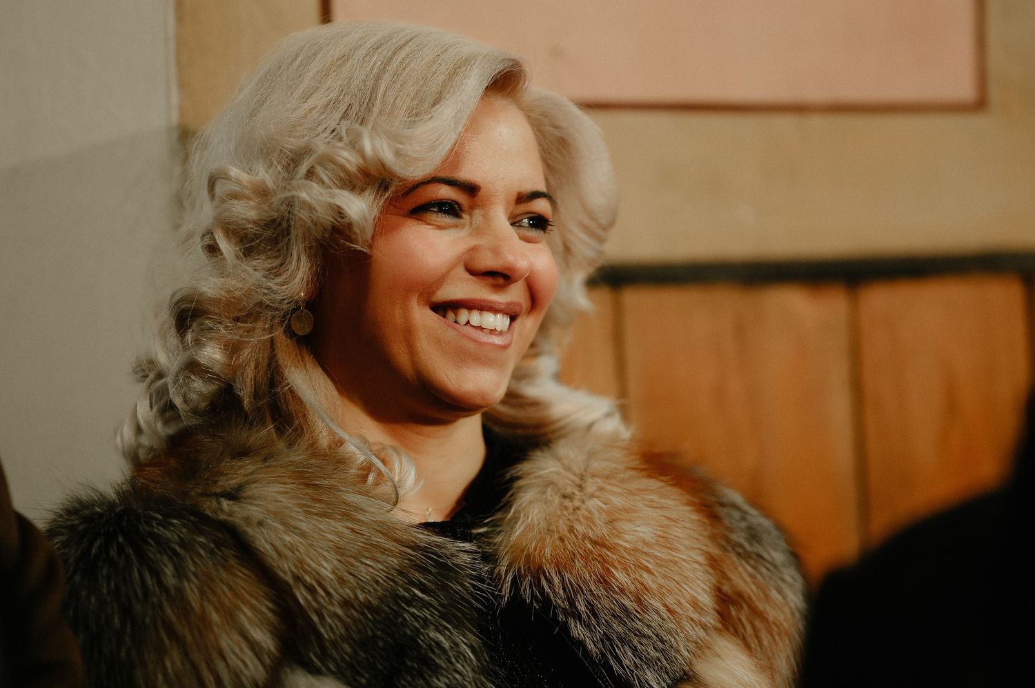 Smiling blonde woman with curly hair wearing a fur coat at a celebration.