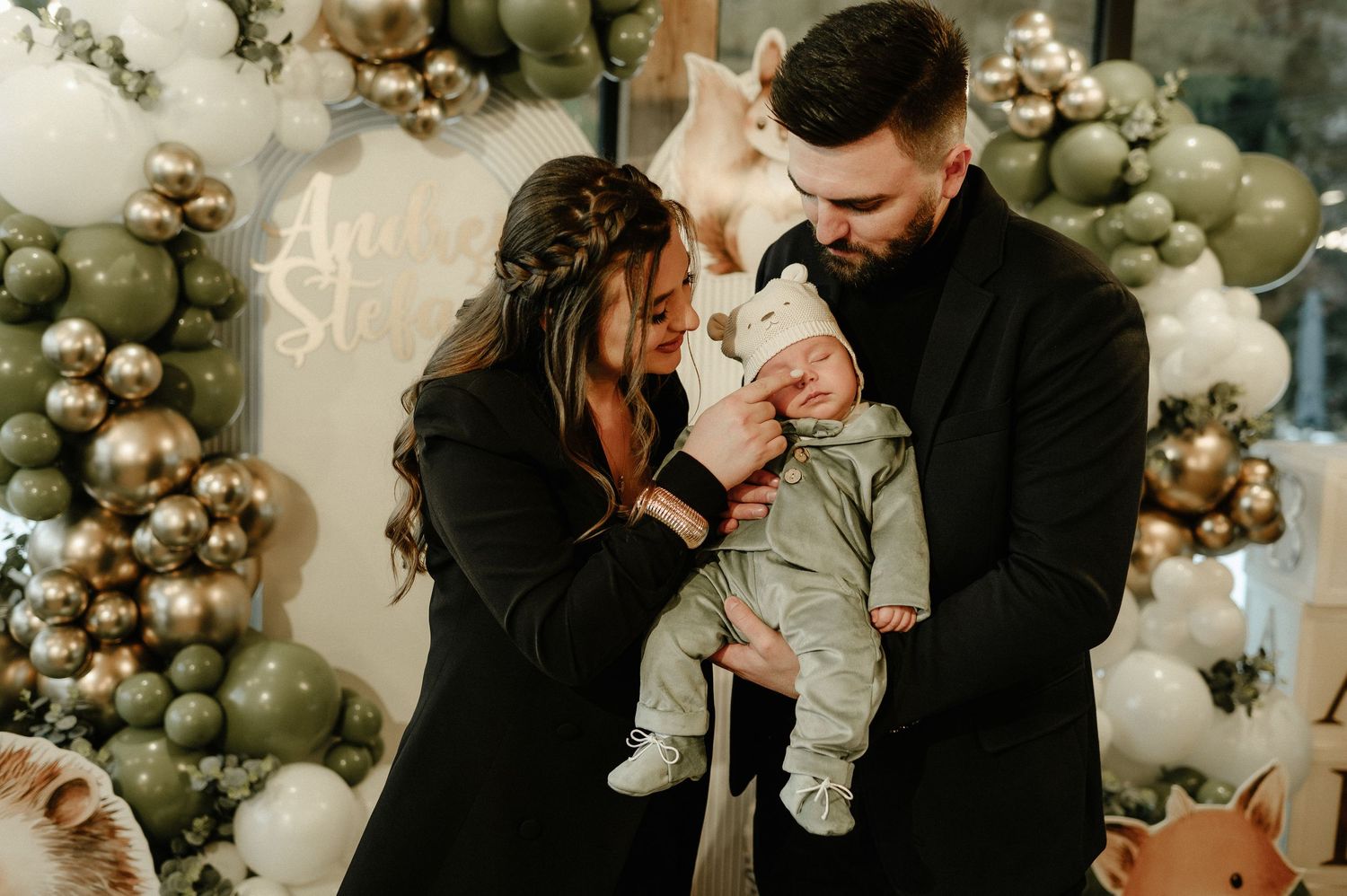 Smiling parents in black outfits holding newborn baby at elegant balloon-decorated baby shower event.