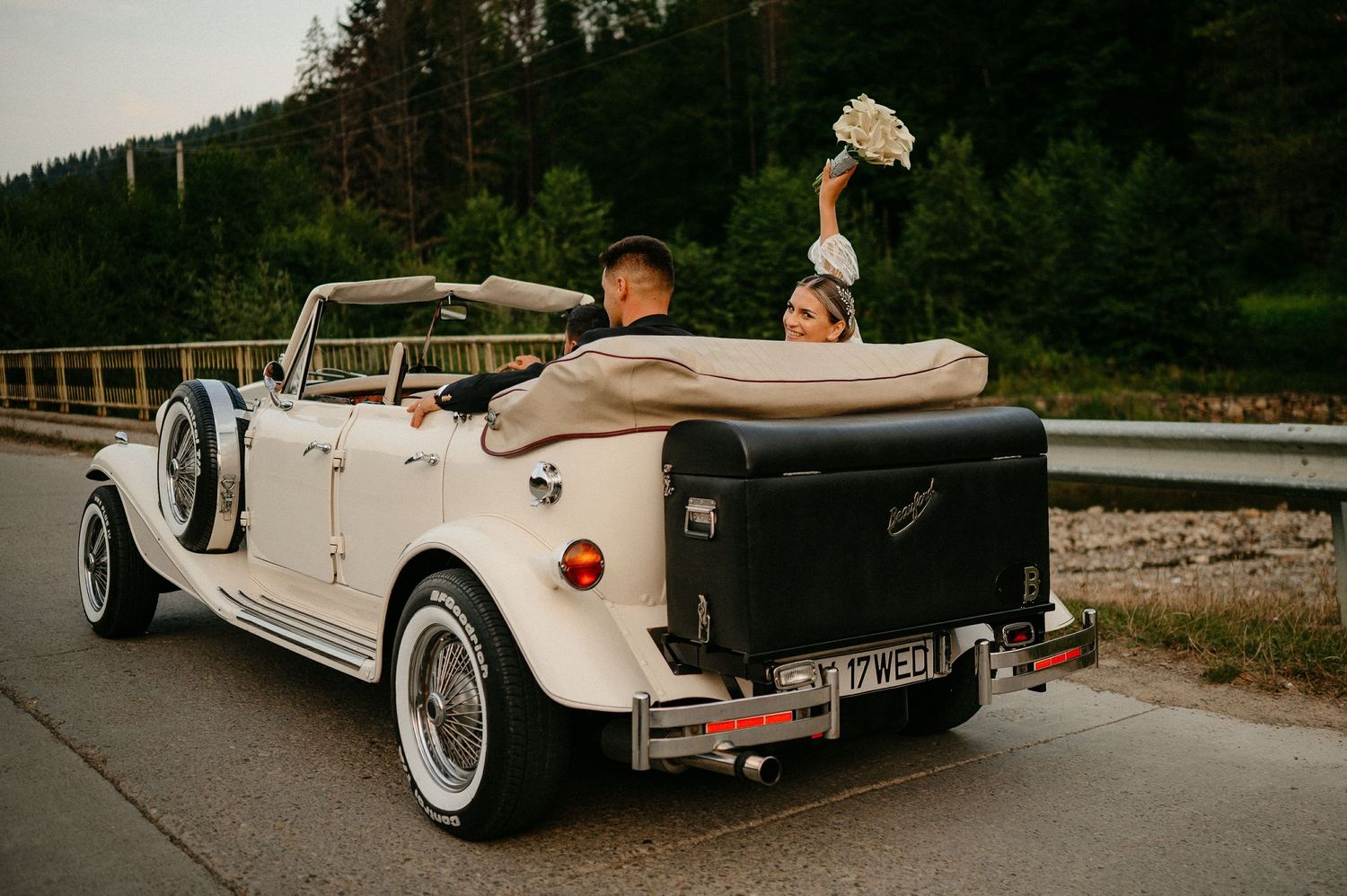 Joyful bride waving bouquet while riding in a classic white vintage convertible wedding car.