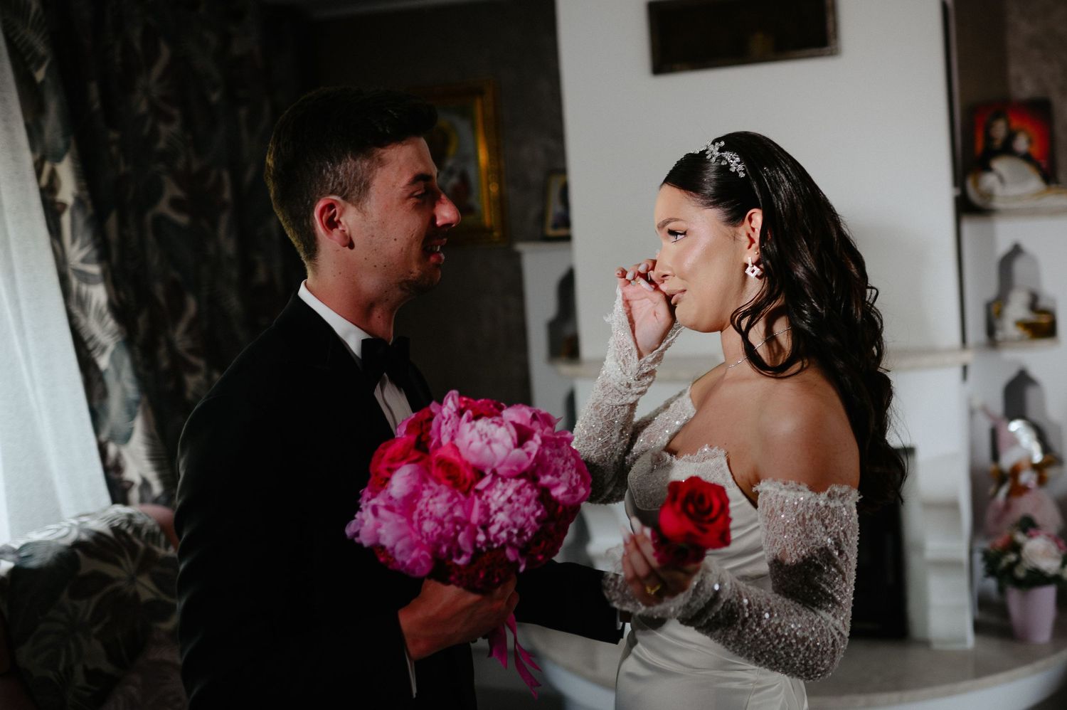 Bride wipes tears while groom tenderly holds her hand, both holding pink peony bouquets at wedding ceremony.