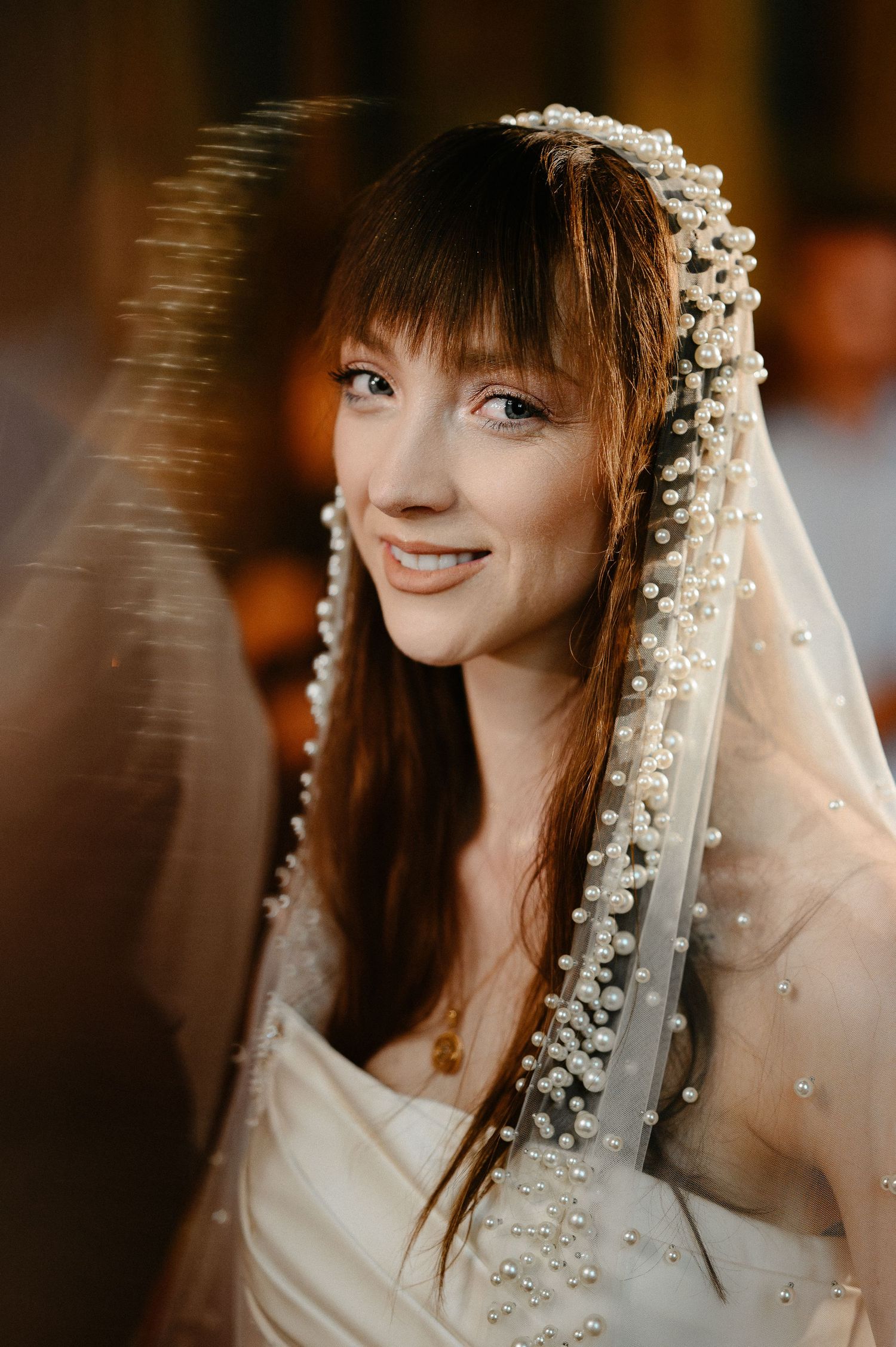 Smiling bride with pearl-embellished veil and bangs glows in warm light at wedding ceremony.