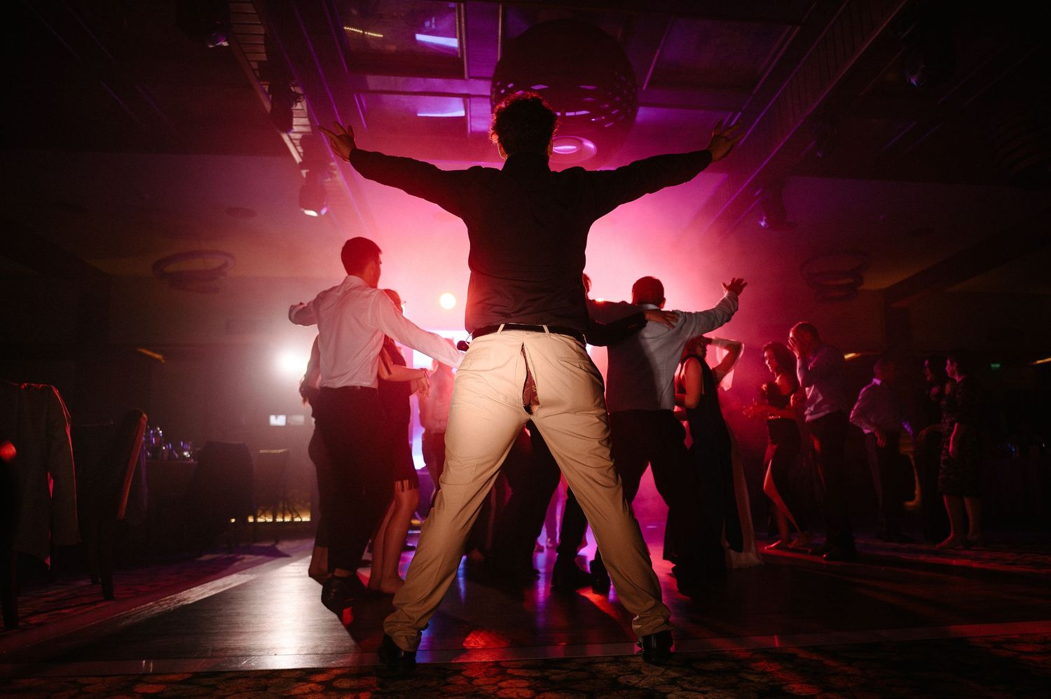 Energetic wedding guests dancing under vibrant pink and red lights on crowded dance floor.