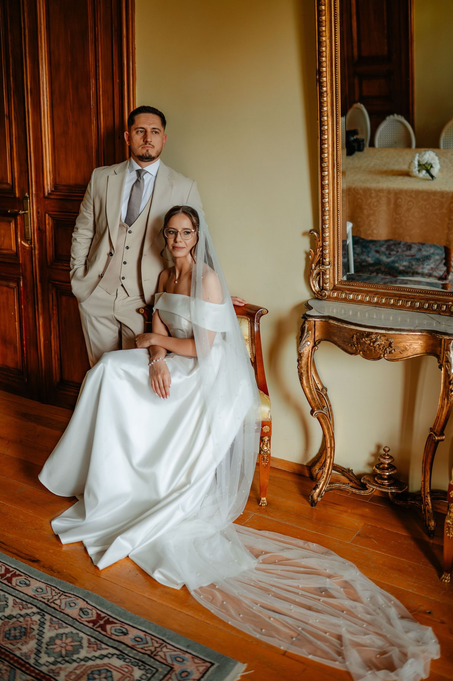 Wedding couple posing in vintage room, bride seated in white gown, groom standing in beige suit.