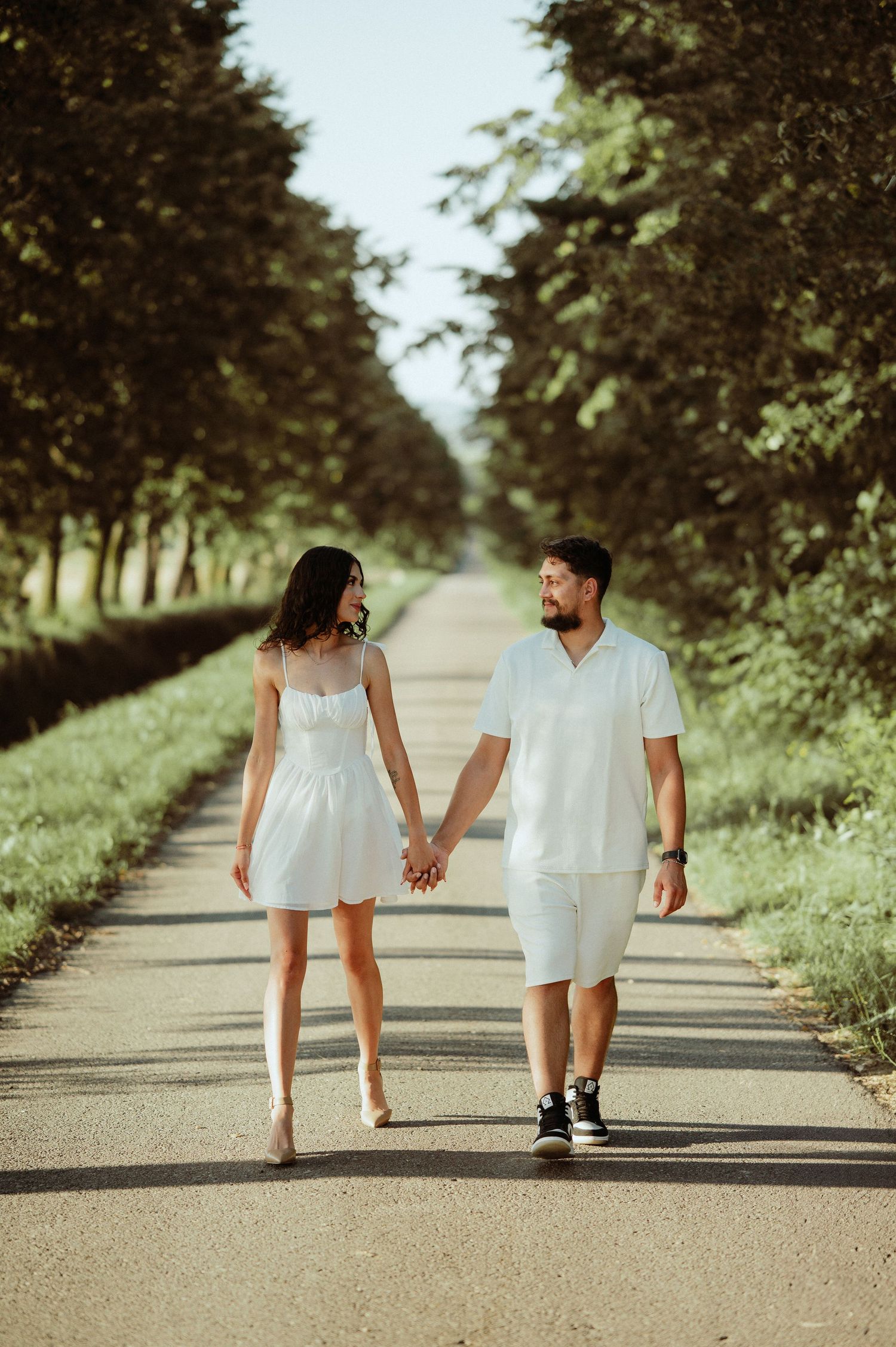 Couple in white outfits holding hands walking down tree-lined road.