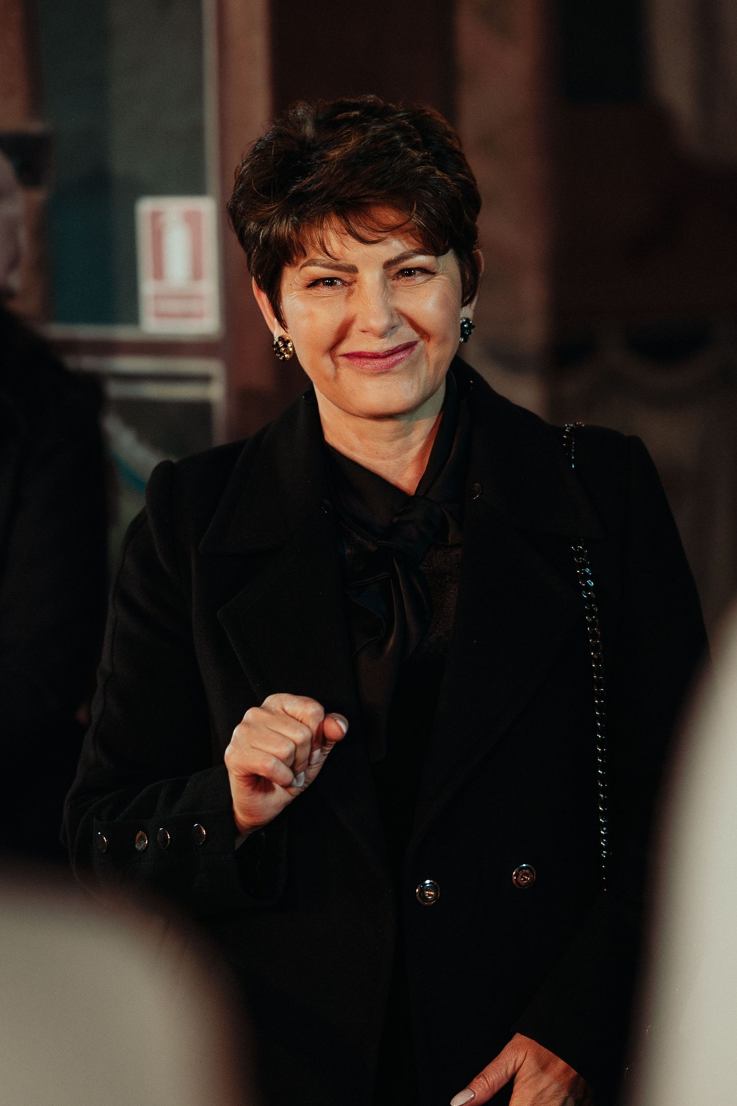 Elegant older woman with short dark hair smiling in a black double-breasted coat.