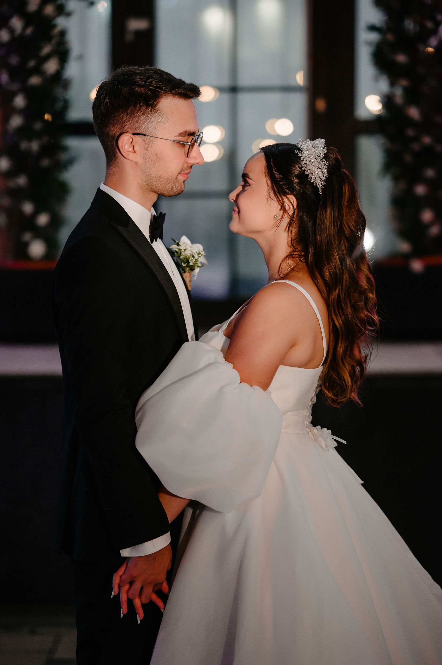 Bride in white off-shoulder gown and groom in black tuxedo share romantic moment at evening wedding venue.