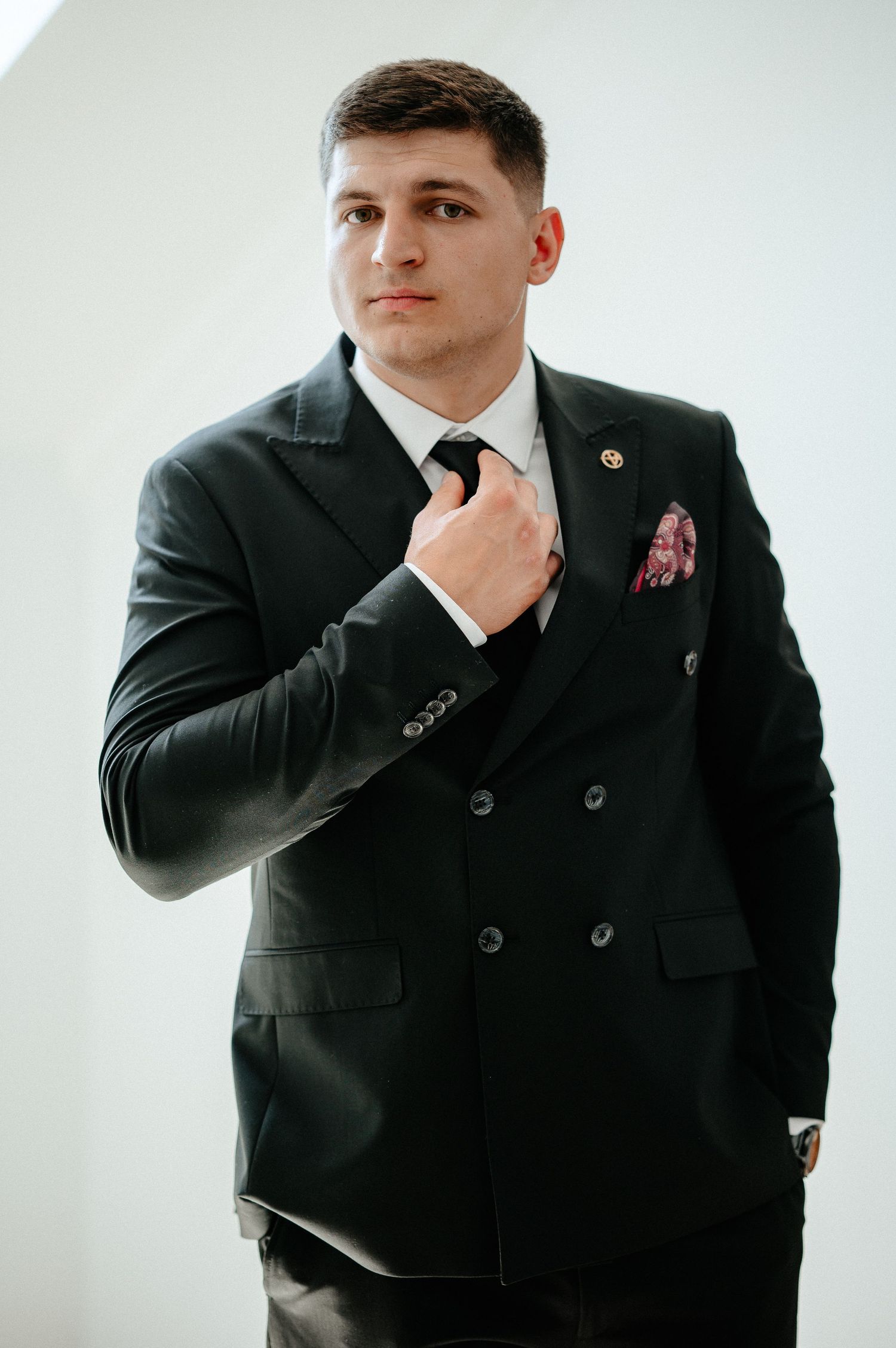 Groom in black double-breasted suit adjusting tie with pink pocket square against white background.