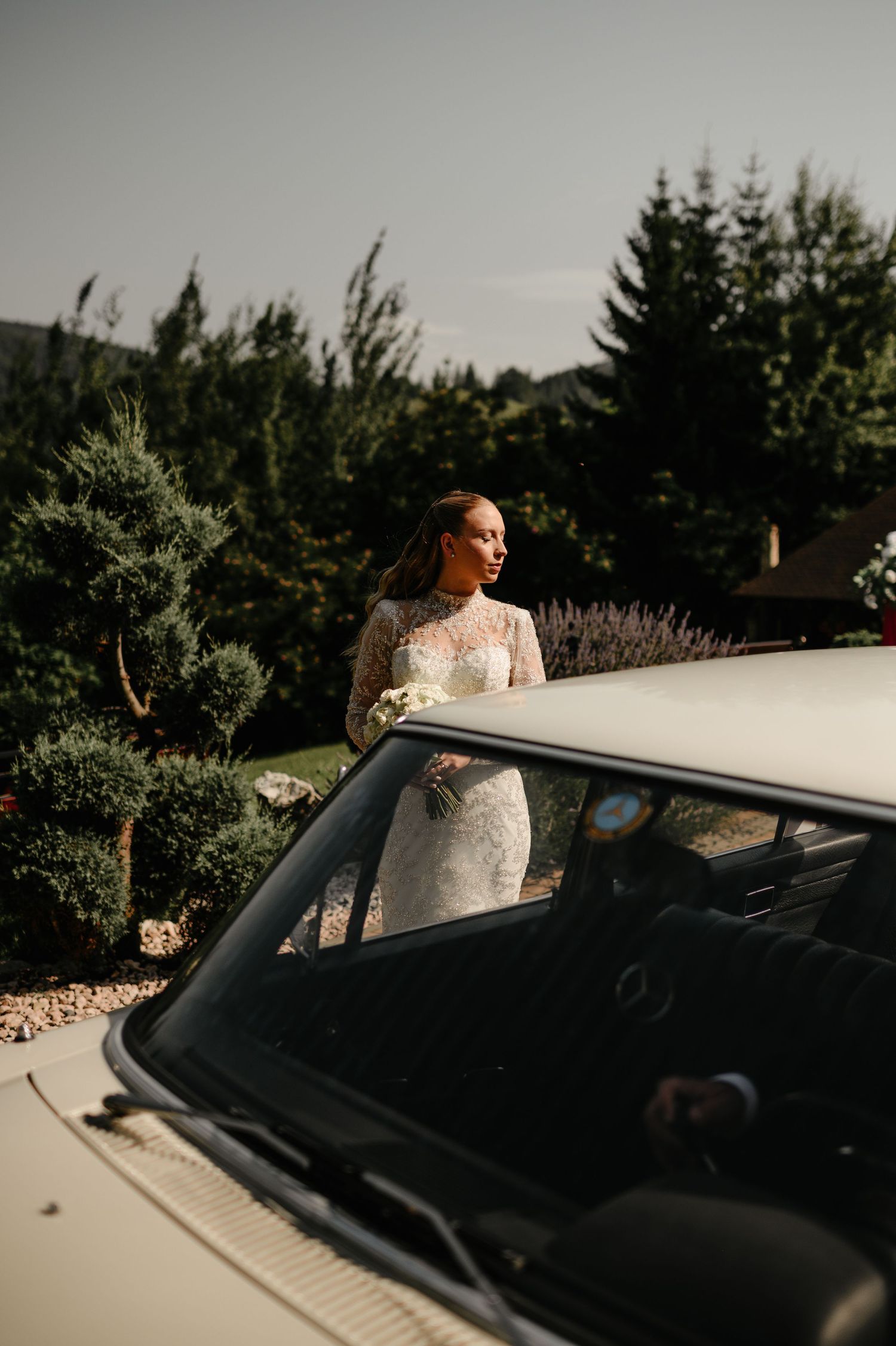 Elegant bride in lace gown holds bouquet beside vintage white Mercedes car outdoors.