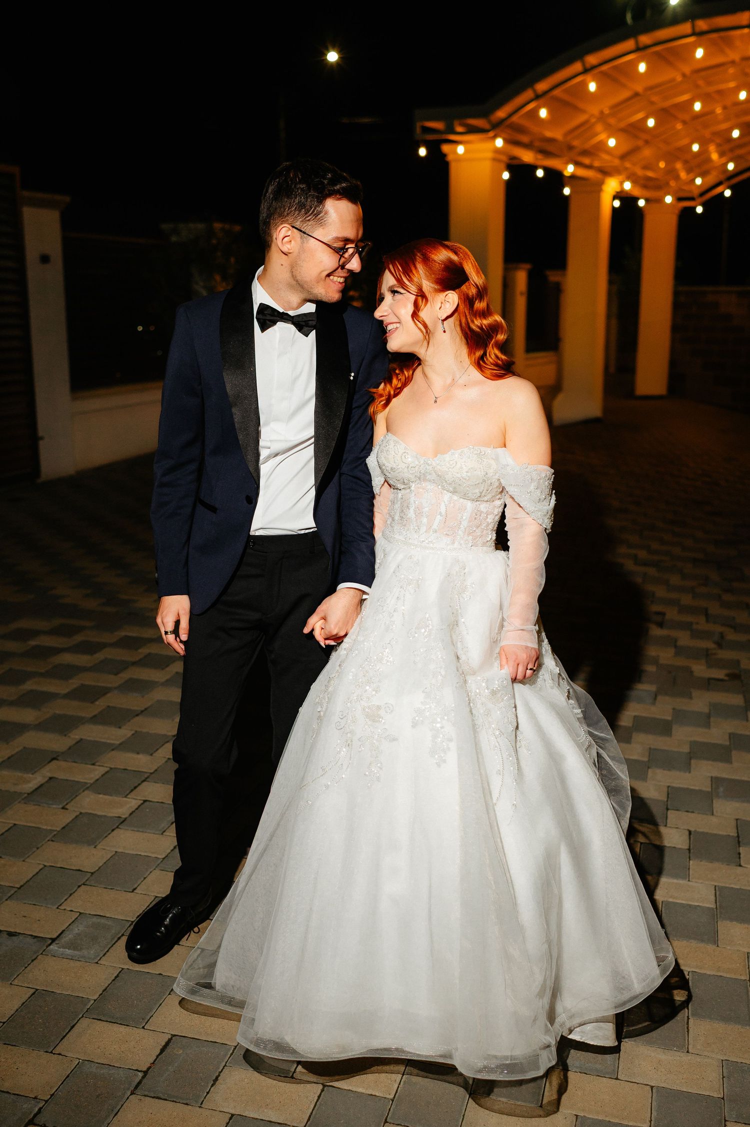 A bride in a white off-shoulder gown and groom in navy tuxedo hold hands smiling at each other at night.