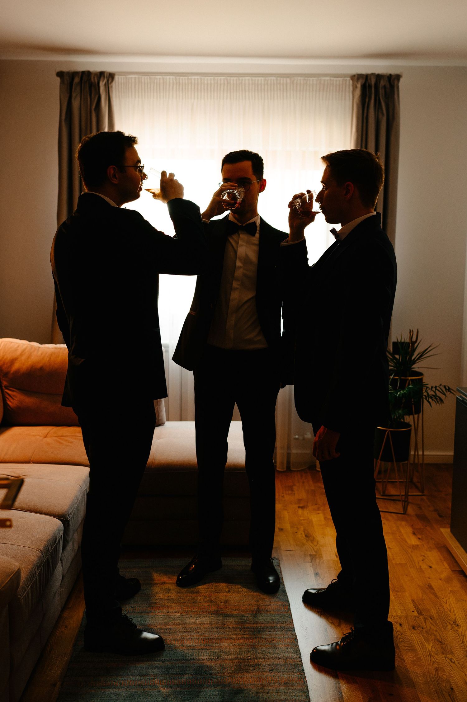 Three groomsmen in suits toasting with shot glasses in a warmly lit room before a wedding ceremony.