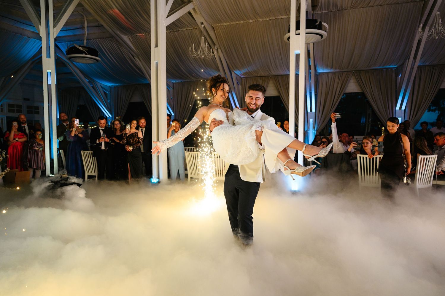 Groom carrying bride on foggy dance floor at elegant wedding reception with blue lighting.