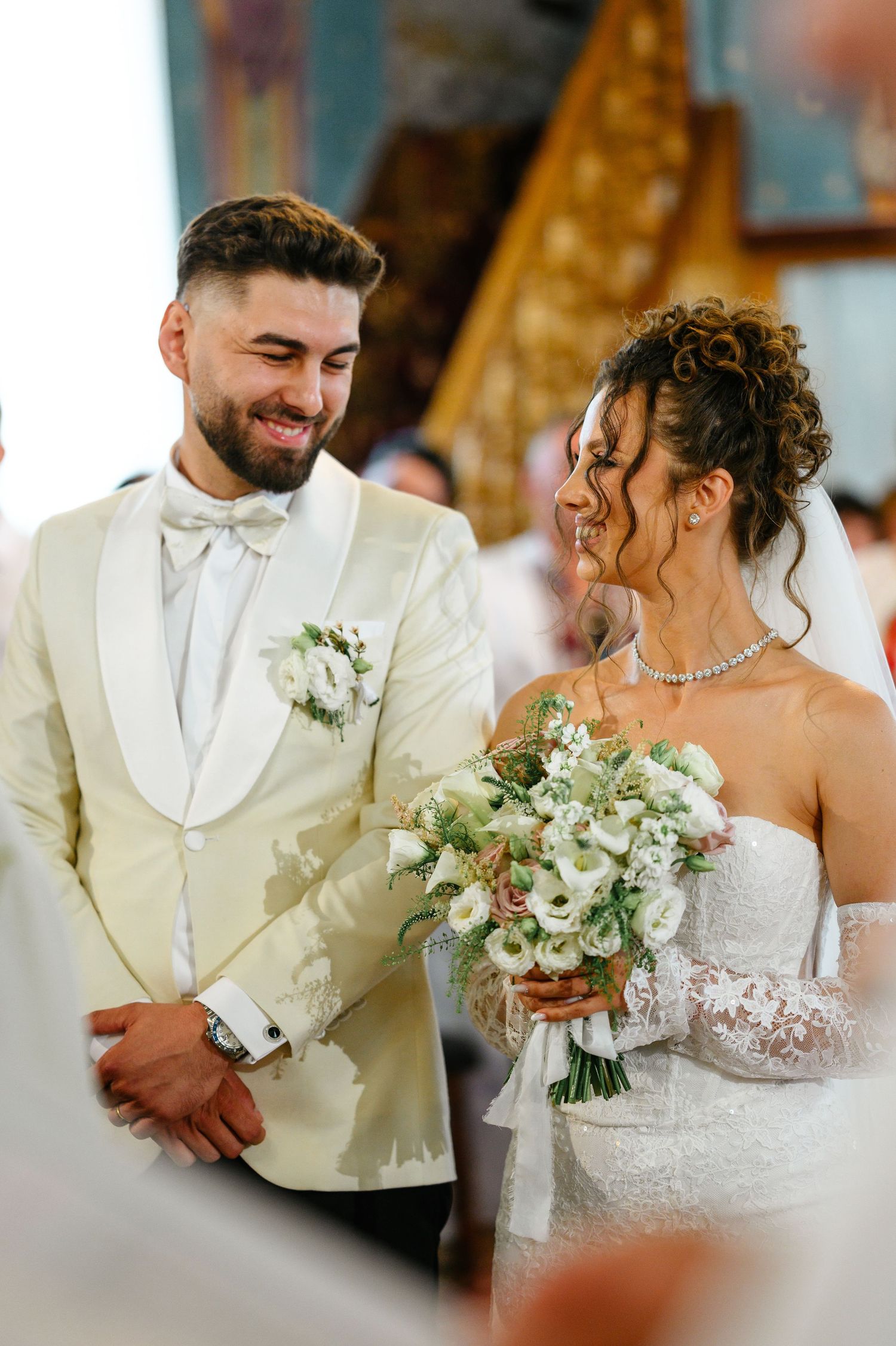 Smiling bride and groom stand at the altar, bride holding white bouquet, groom in cream tuxedo with boutonniere.