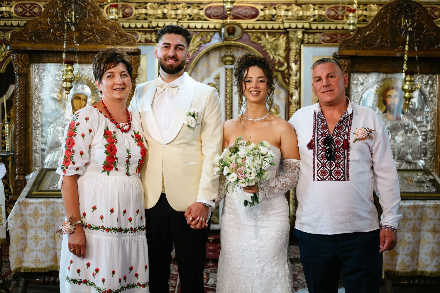 Wedding party of four posing in ornate church, bride in white gown holding bouquet, groom in cream suit, parents in traditional attire.