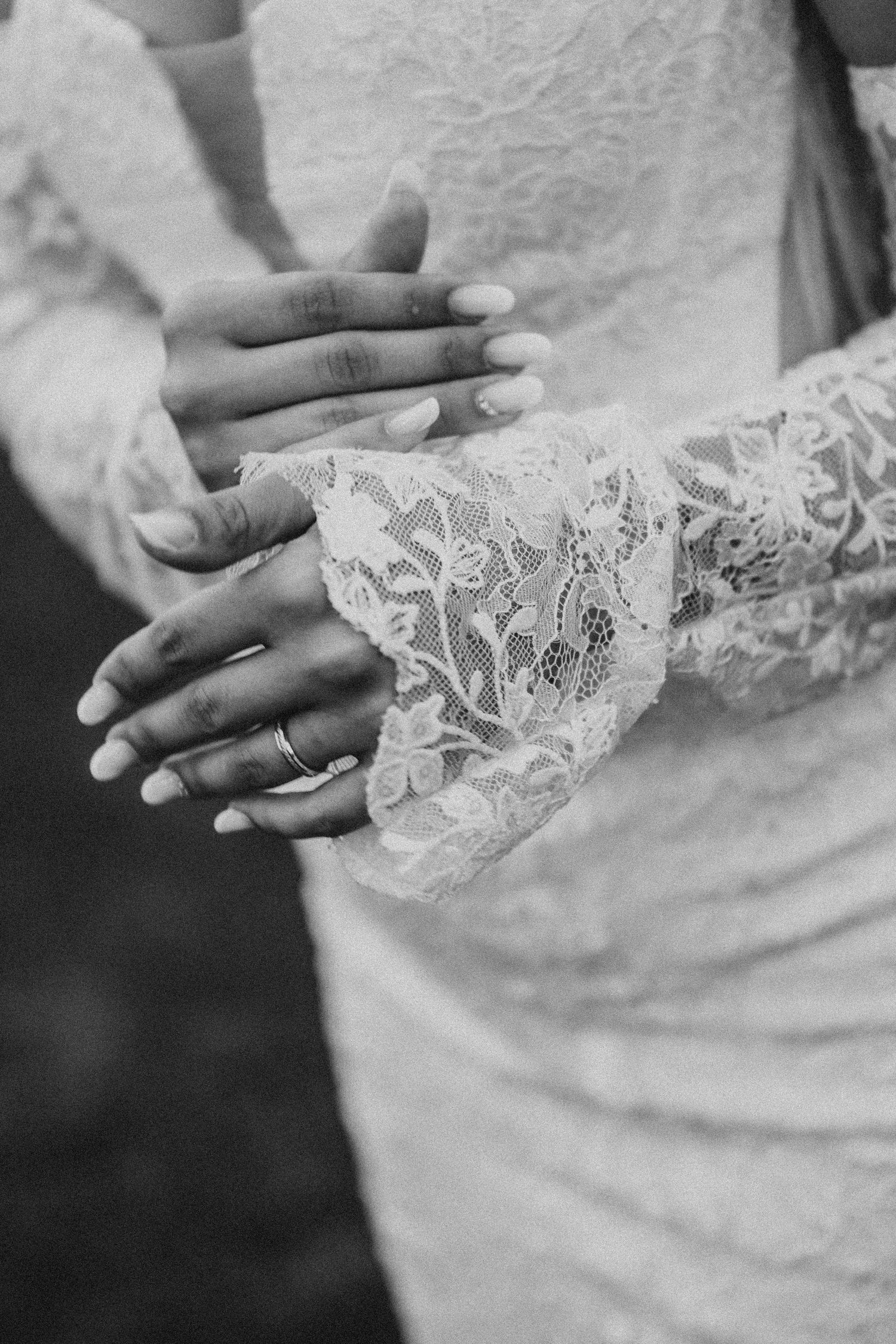 Black and white photo of bride and groom hands clasped together, wedding ring visible on lace sleeve dress.