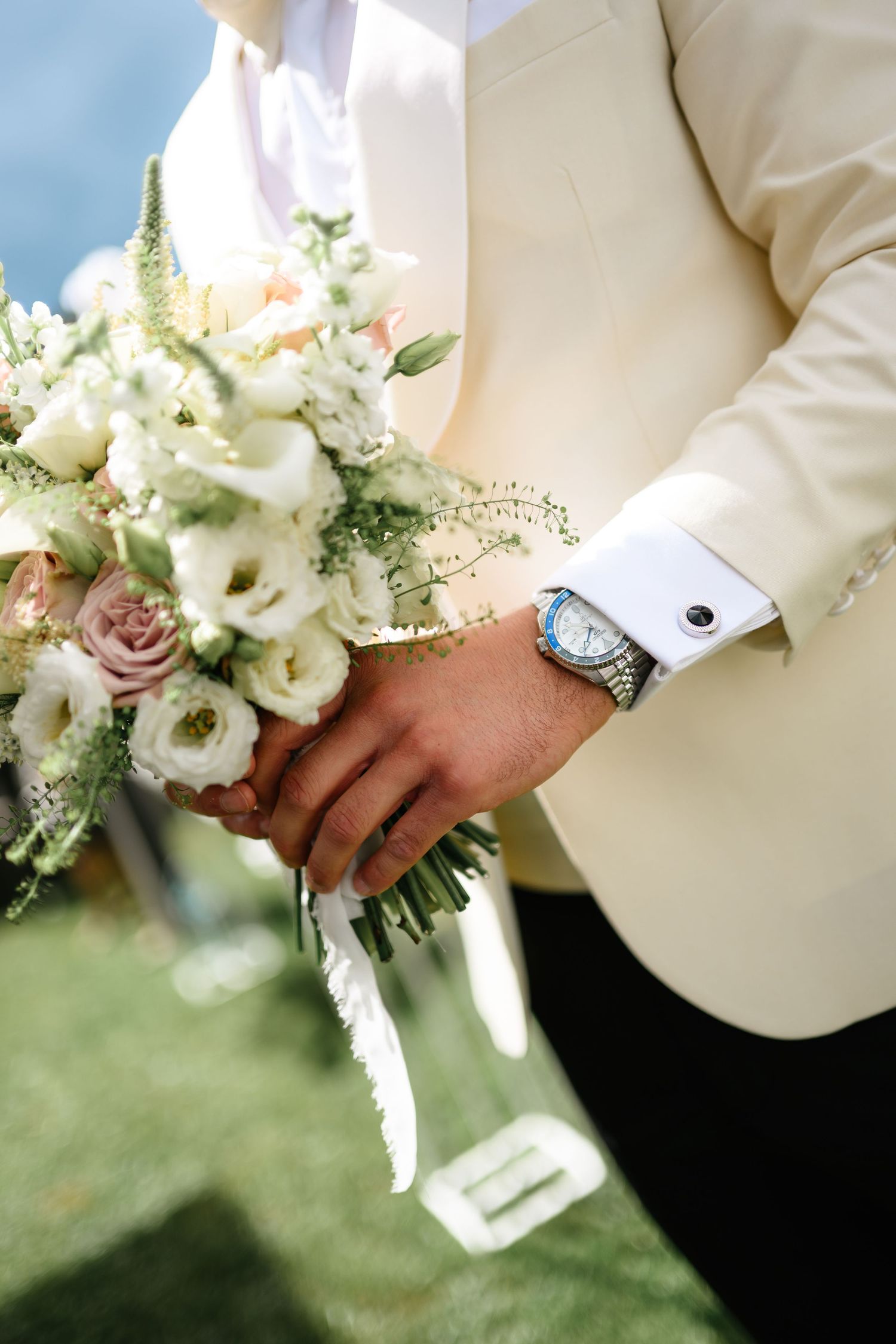 Groom in ivory tuxedo holding elegant white and blush wedding bouquet with cufflinks and silver watch visible.