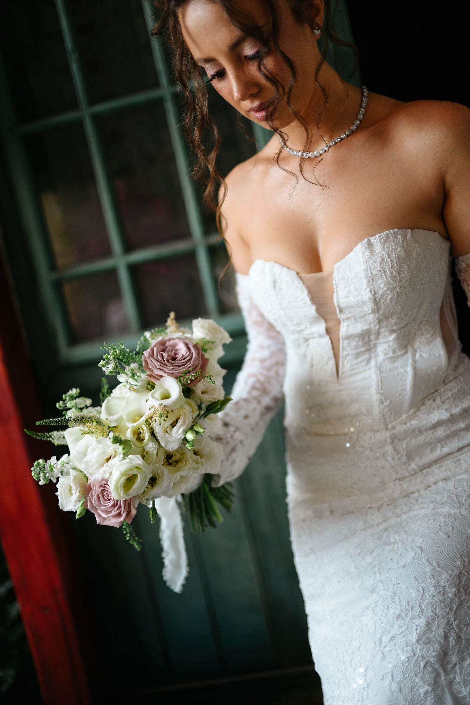 Bride in strapless lace gown holding bouquet of white and mauve roses with pearl necklace.