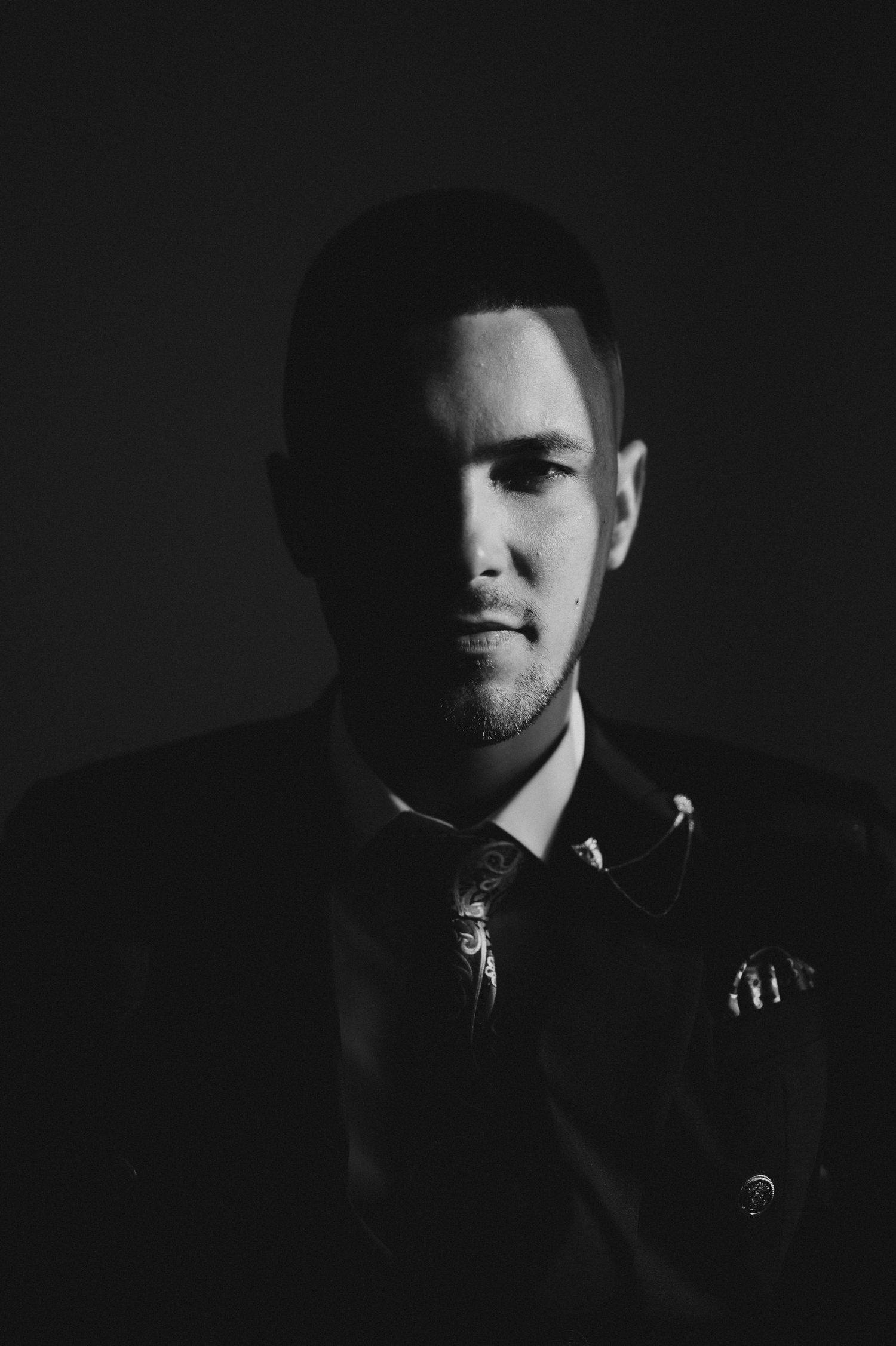 Black and white dramatic portrait of a man in a suit and tie with moody lighting against a dark background.