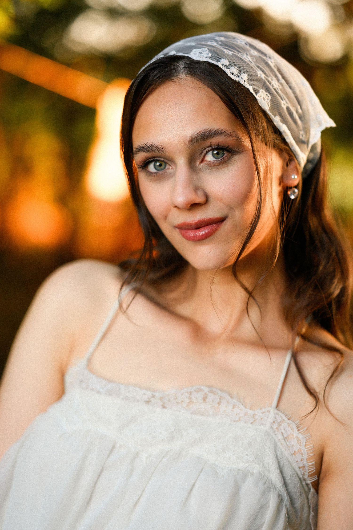 Young woman with blue eyes wearing white lace dress and headscarf smiling outdoors at golden hour.