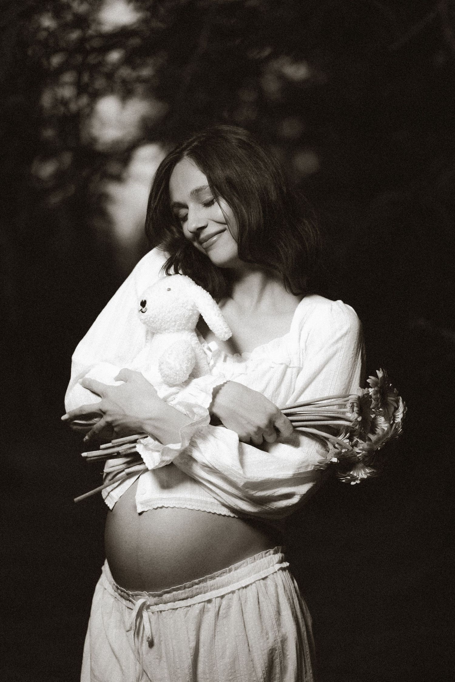 Black and white photo of smiling pregnant woman holding stuffed bunny and flowers outdoors.