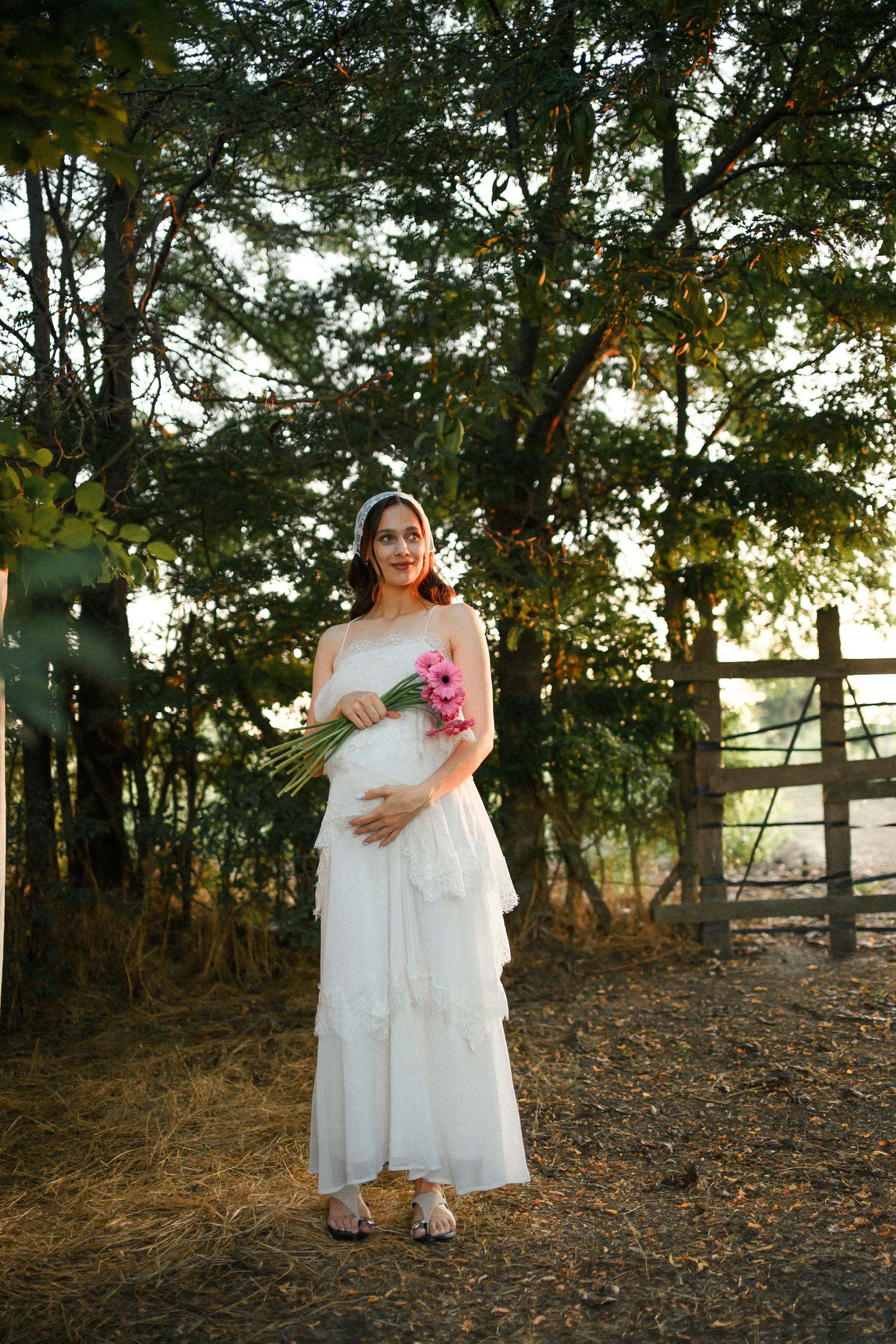 Pregnant woman in white dress holding pink flowers outdoors near wooden fence and trees in golden hour light.