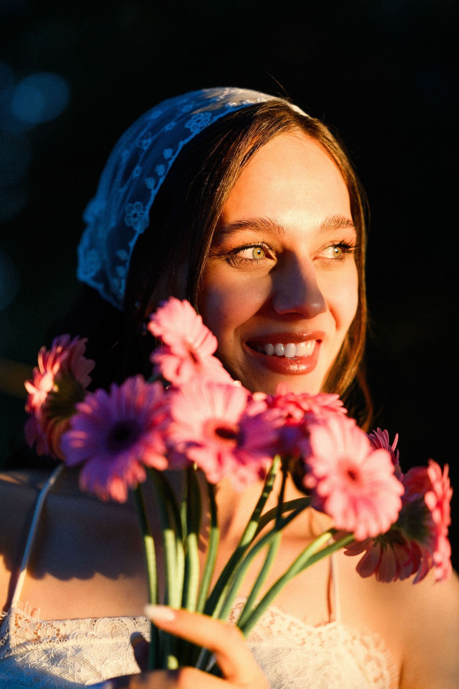 Smiling young woman holding pink gerbera daisies, wearing a white lace top and blue headscarf in golden sunlight.