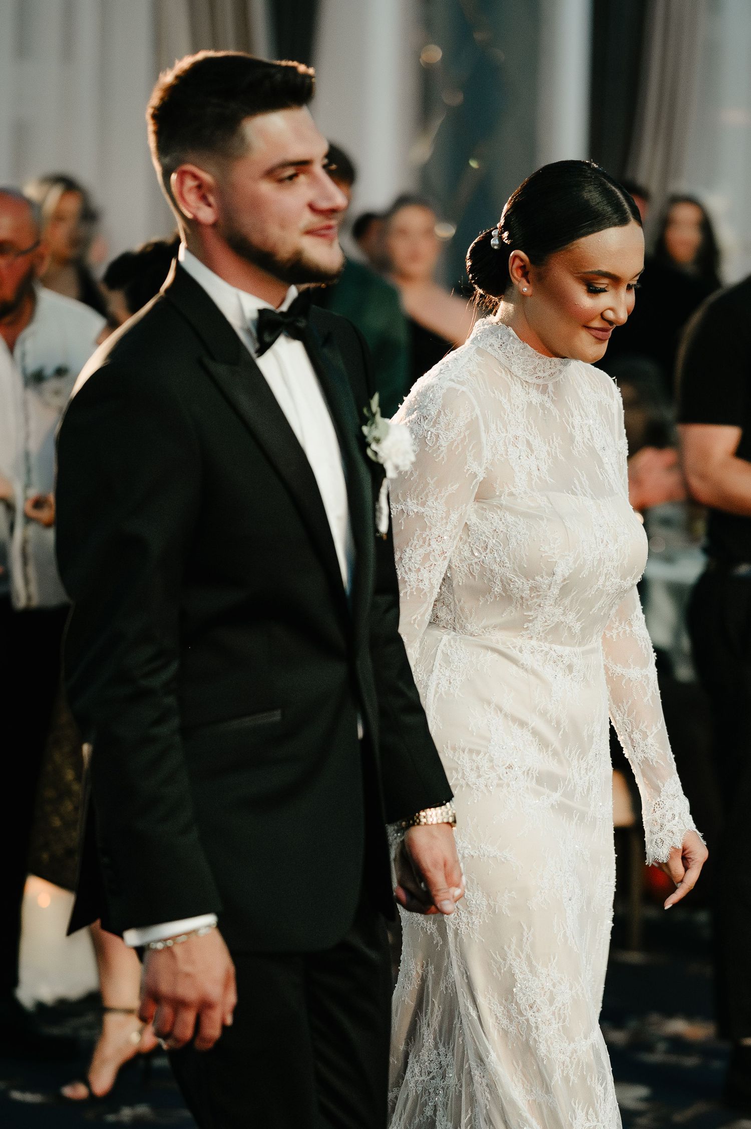 Bride in lace gown and groom in black tuxedo walking hand in hand at wedding reception.