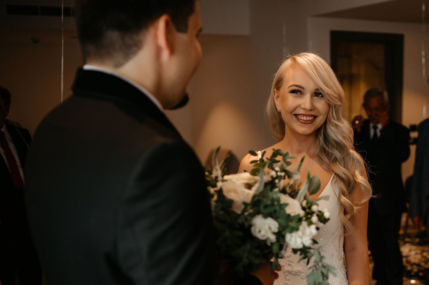 Radiant blonde bride smiling at groom while holding white rose bouquet at indoor wedding venue.