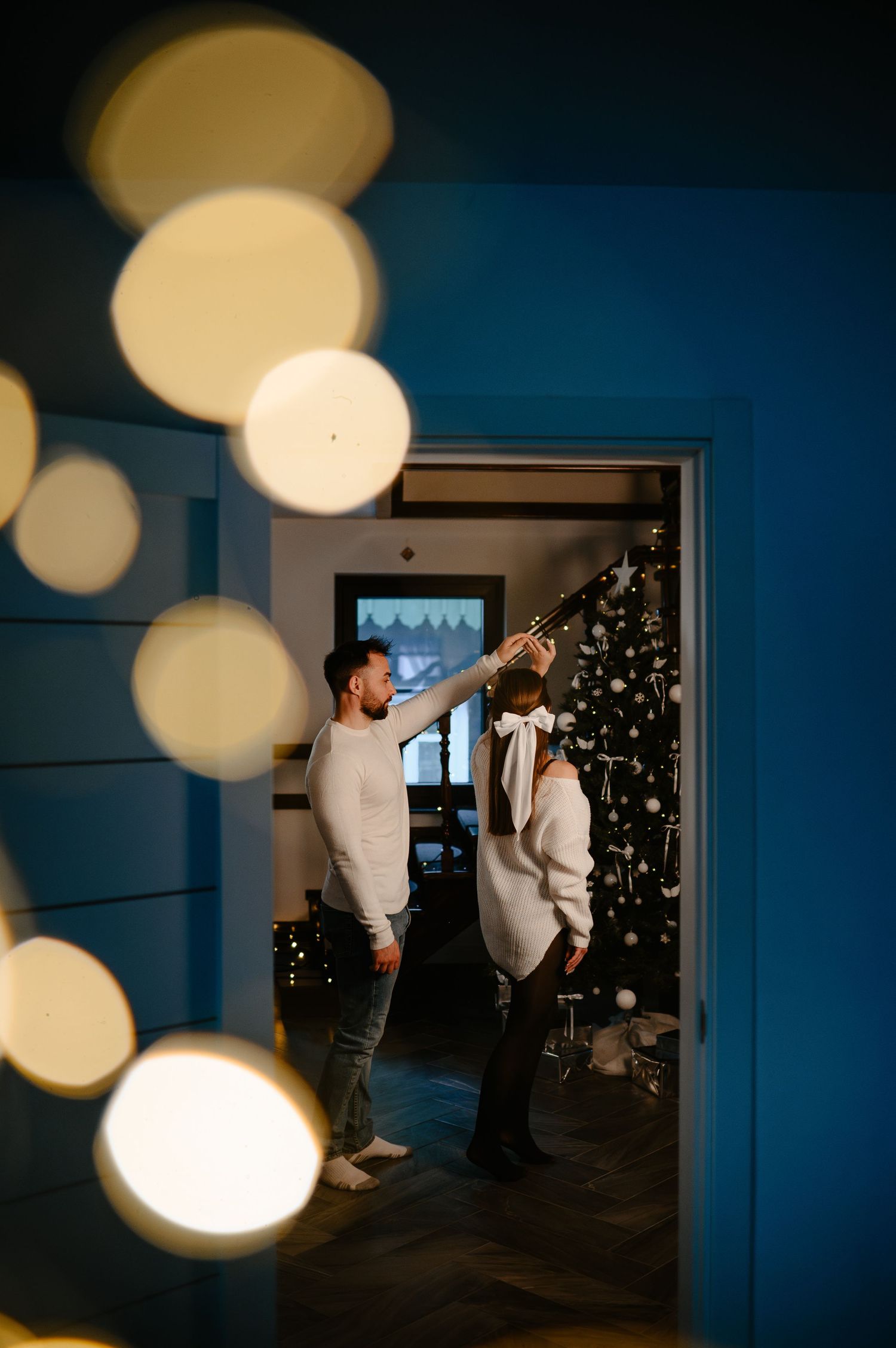 Couple decorating a white Christmas tree in a blue-walled room with glowing bokeh lights.