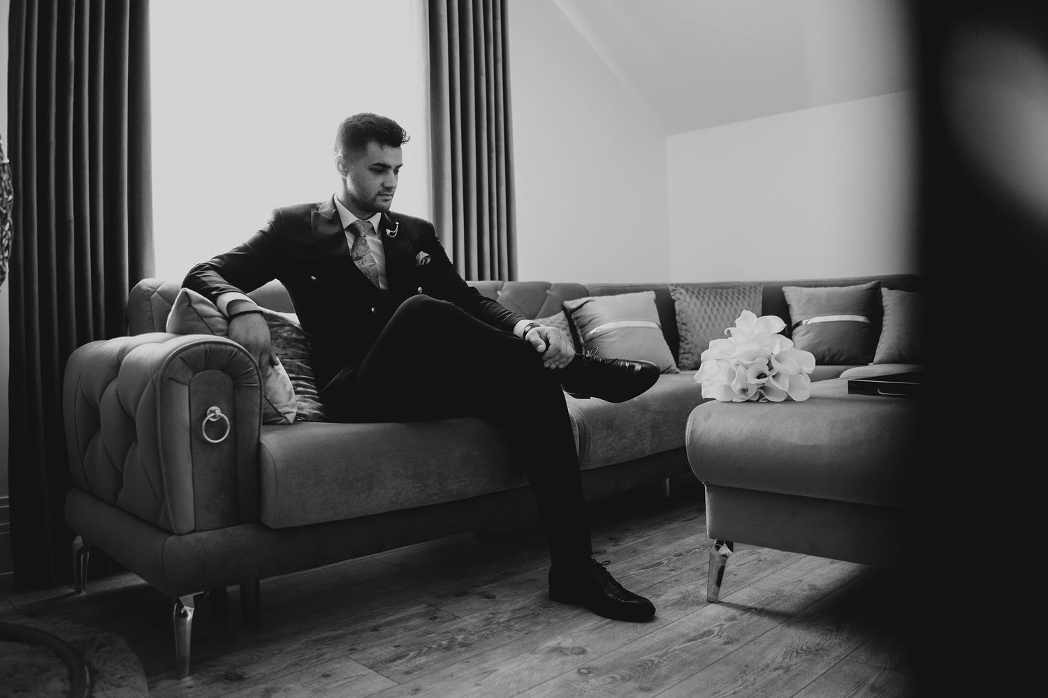 Groom in black tuxedo sitting on sofa with wedding bouquet nearby.