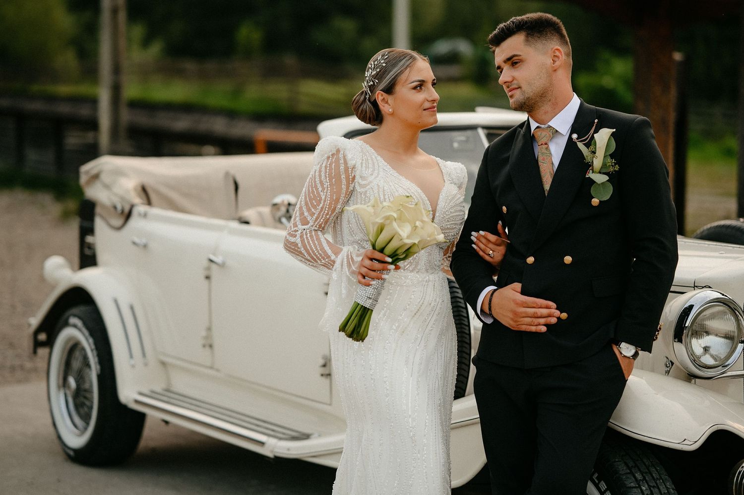 Newlywed couple sharing a loving glance beside a white vintage convertible wedding car.