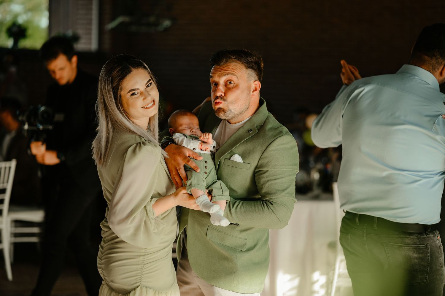 Smiling couple in green outfits holding their newborn baby at a bright indoor celebration event.
