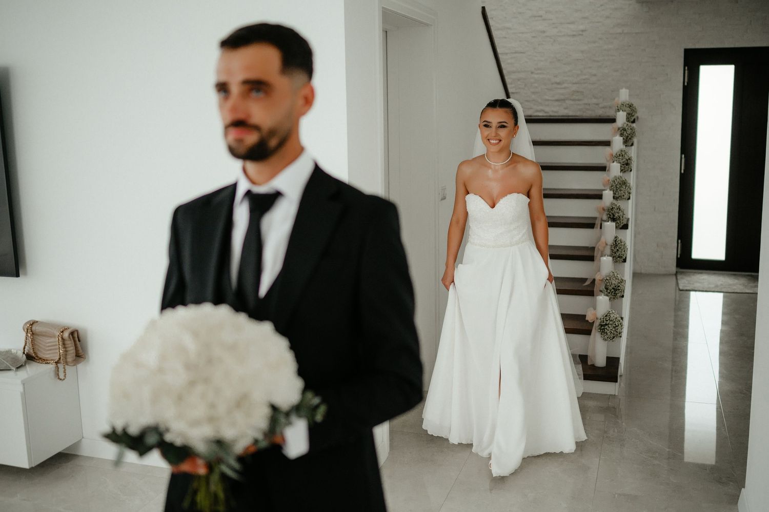 Bride in strapless gown walks toward groom holding white floral bouquet during wedding first look.