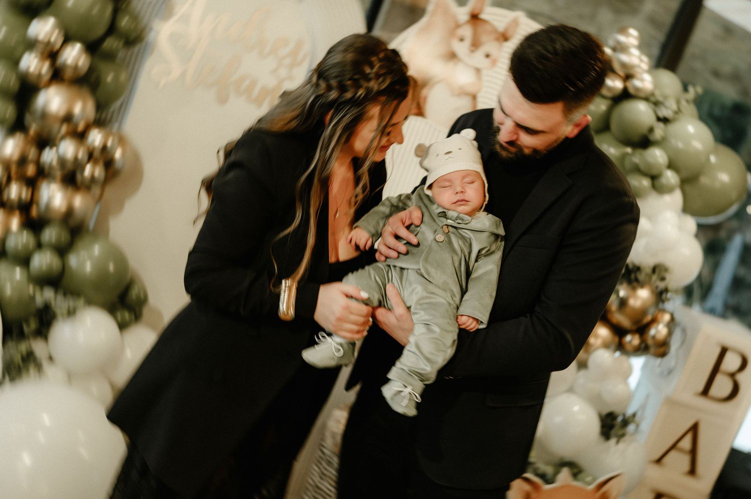 Parents in black outfits lovingly looking at newborn baby at festive balloon-decorated baby shower.
