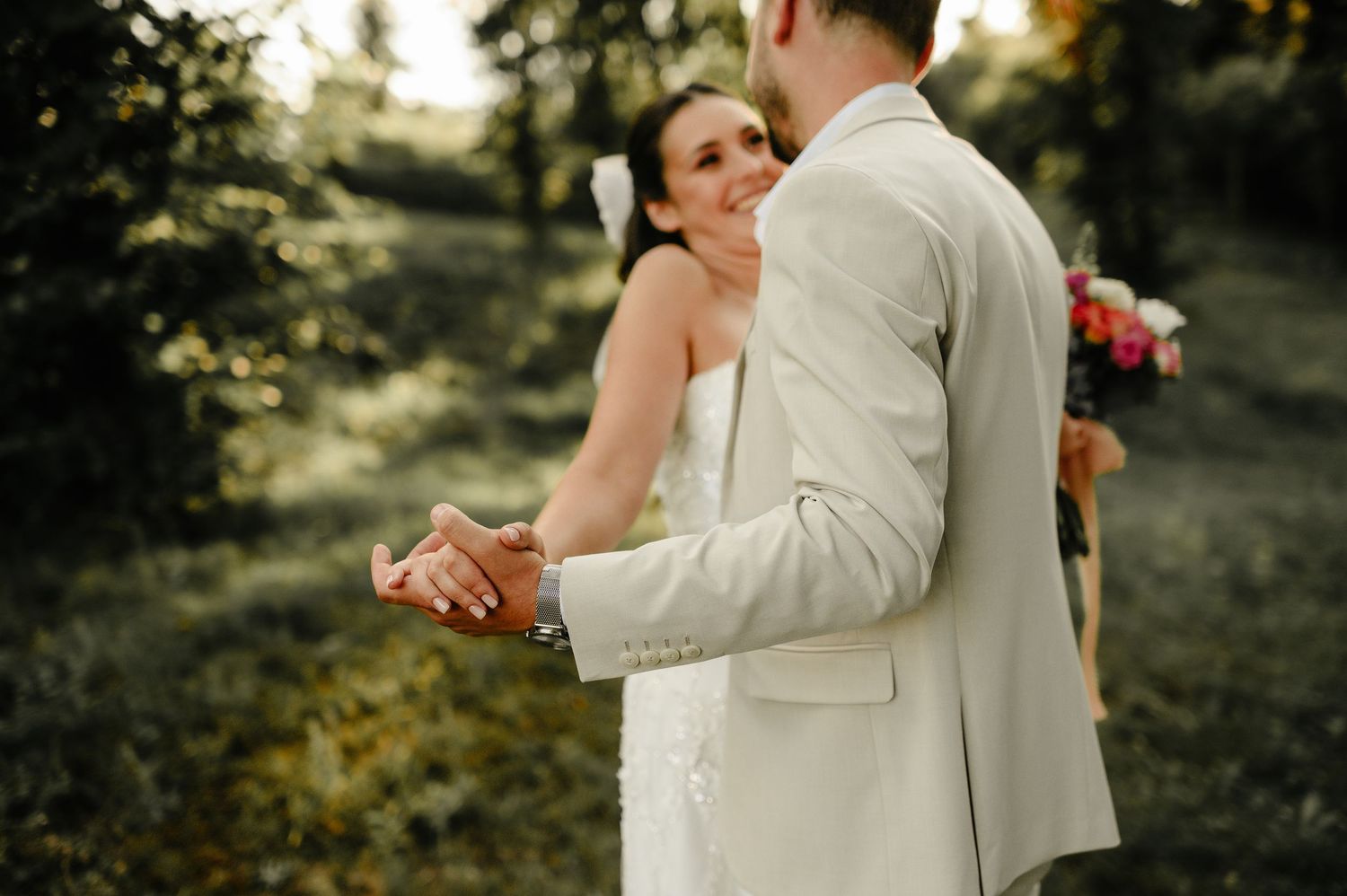 Emotional bride smiling while holding grooms hands in lush green outdoor wedding ceremony setting.