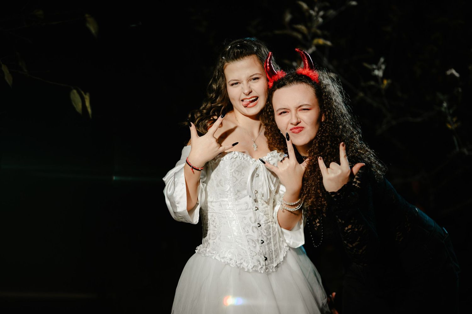 Two young women in Halloween costumes posing together, one in white bridal gown, one with devil horns.
