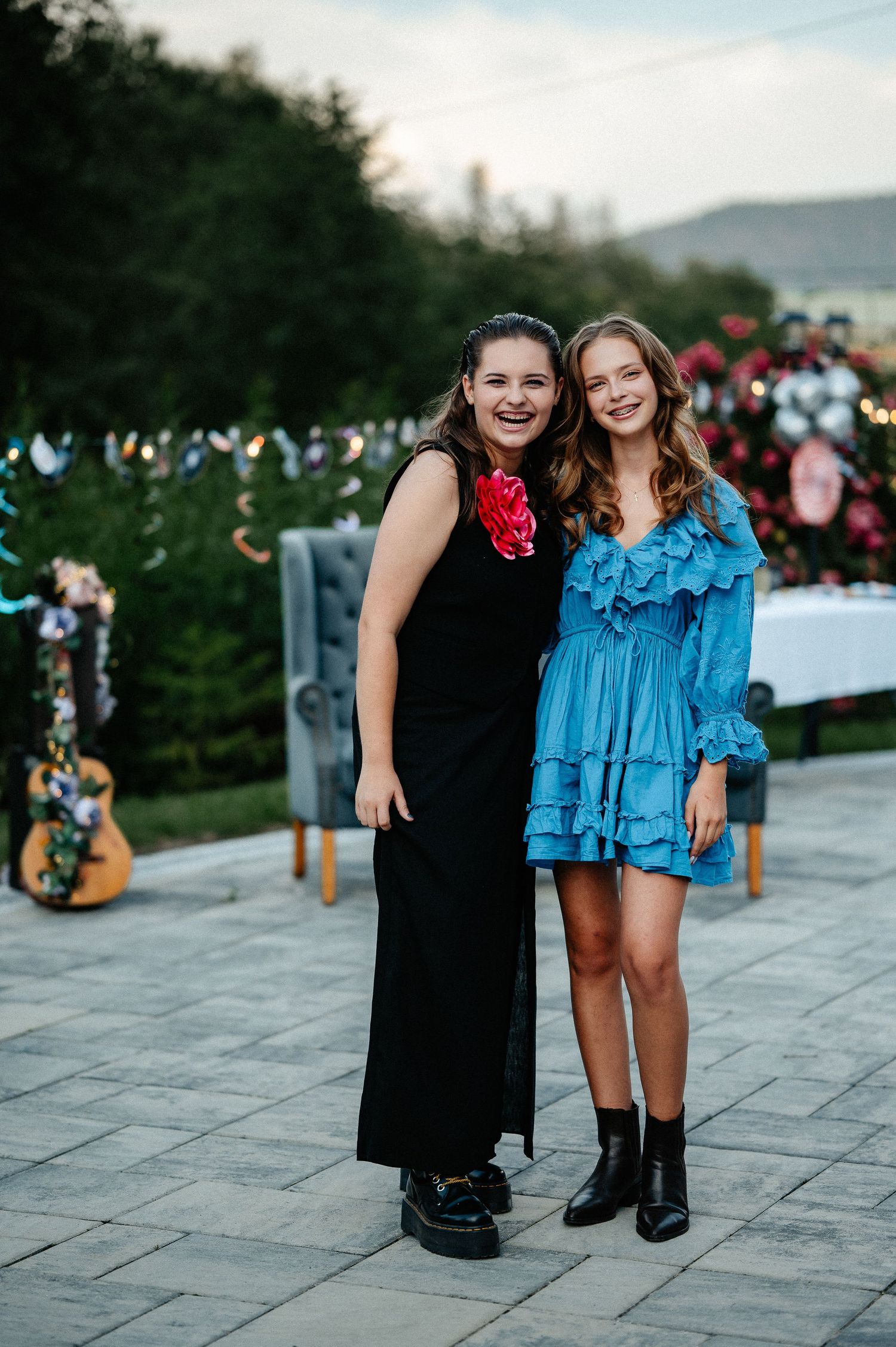 Two young women smiling together outdoors, one in a black maxi dress and one in a blue ruffled mini dress.
