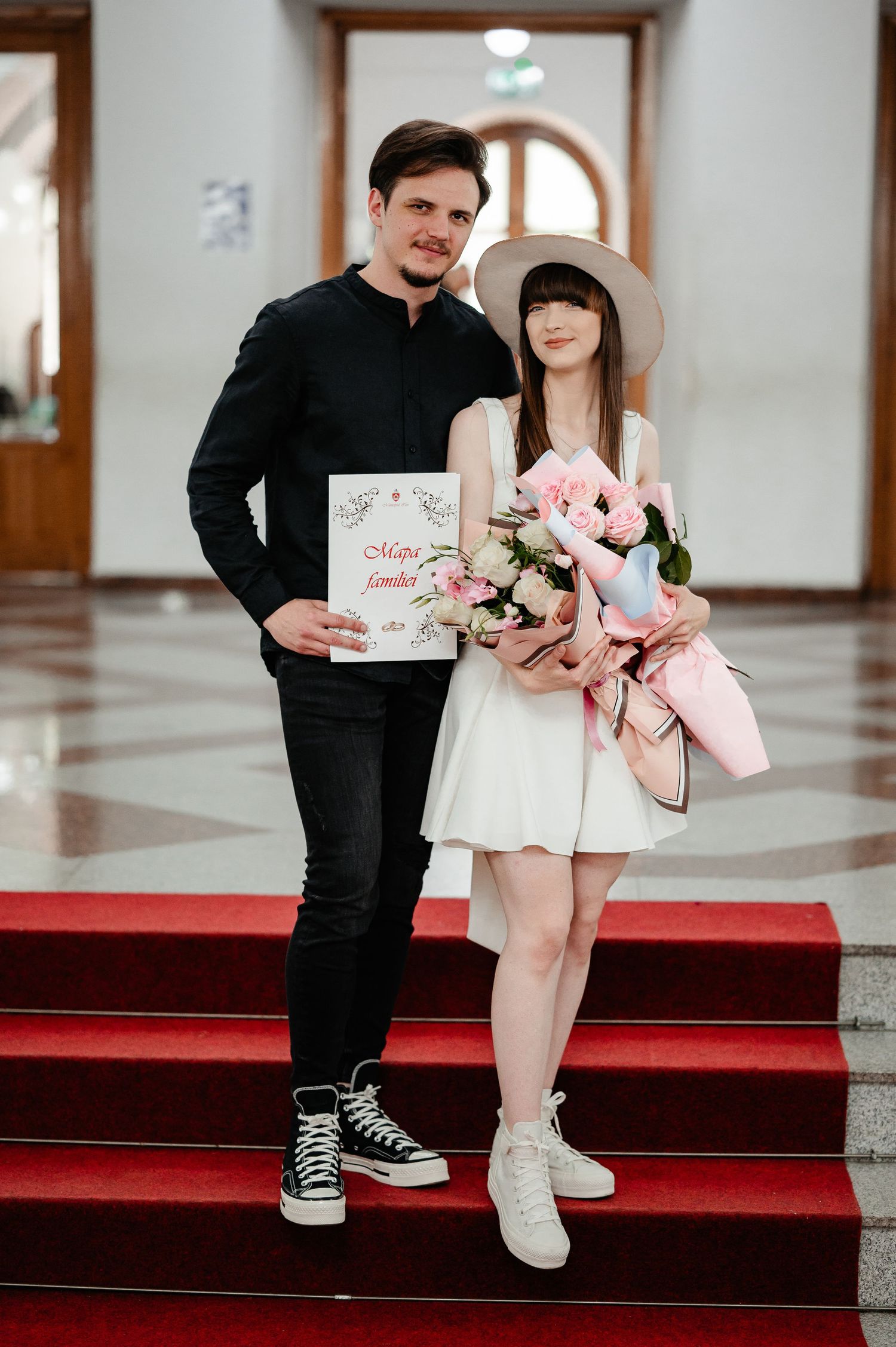 Newlyweds pose on red-carpeted stairs, groom holding marriage certificate, bride with pink flower bouquet and white hat.
