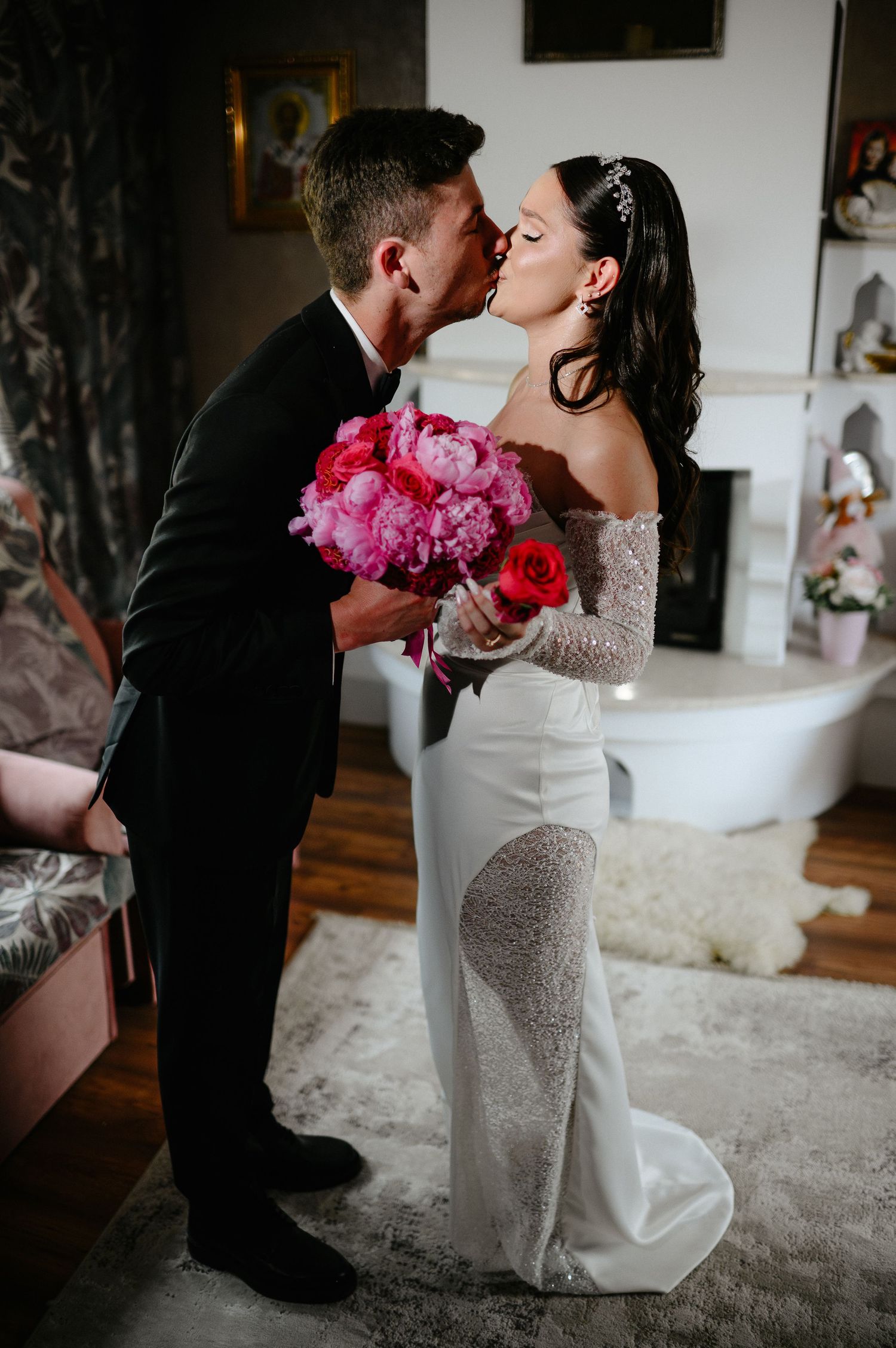 Bride and groom share a romantic kiss, bride holding vibrant pink peony bouquet, wearing elegant lace wedding gown.
