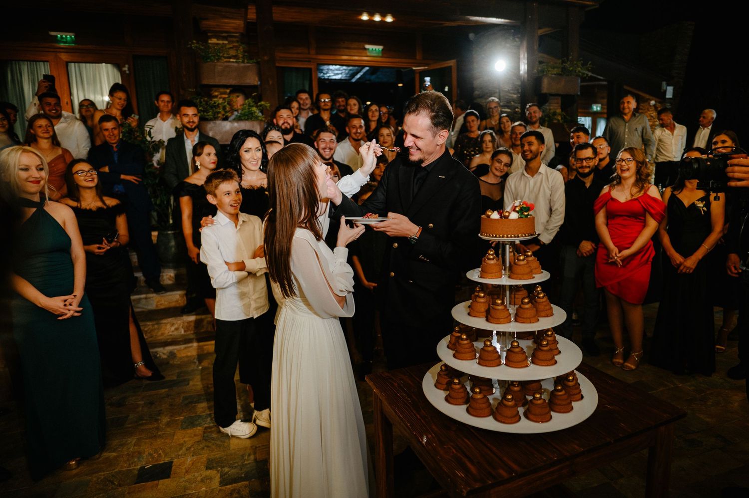 Wedding couple cuts tiered cake display while guests watch at elegant evening reception.