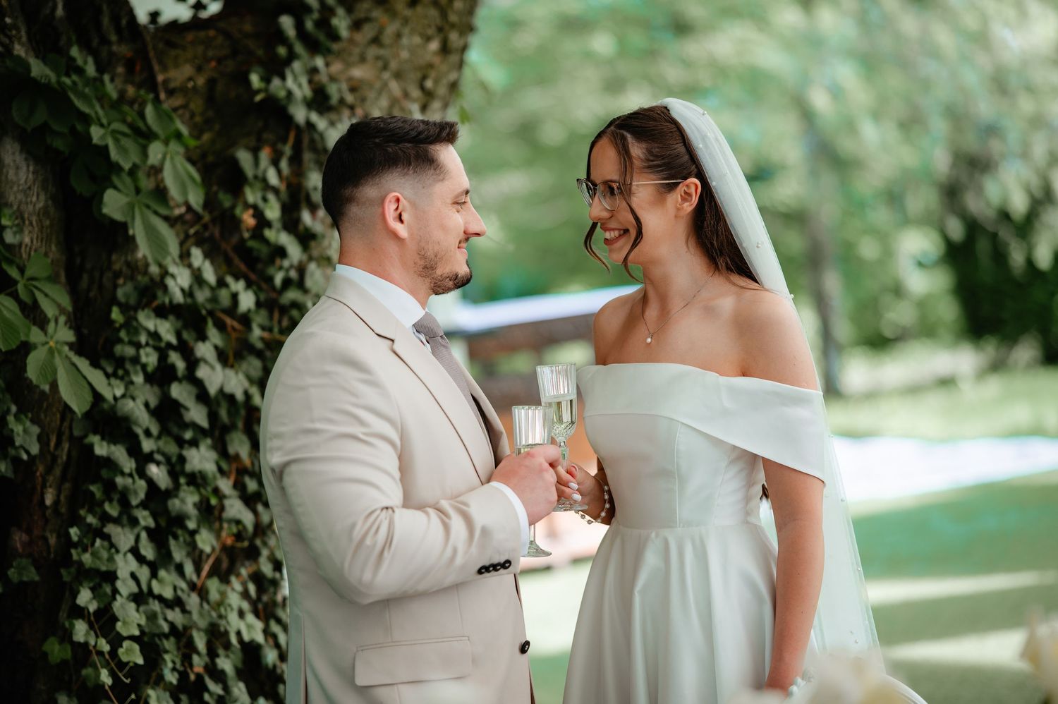 Bride and groom toasting with champagne glasses outdoors near ivy-covered tree on their wedding day.