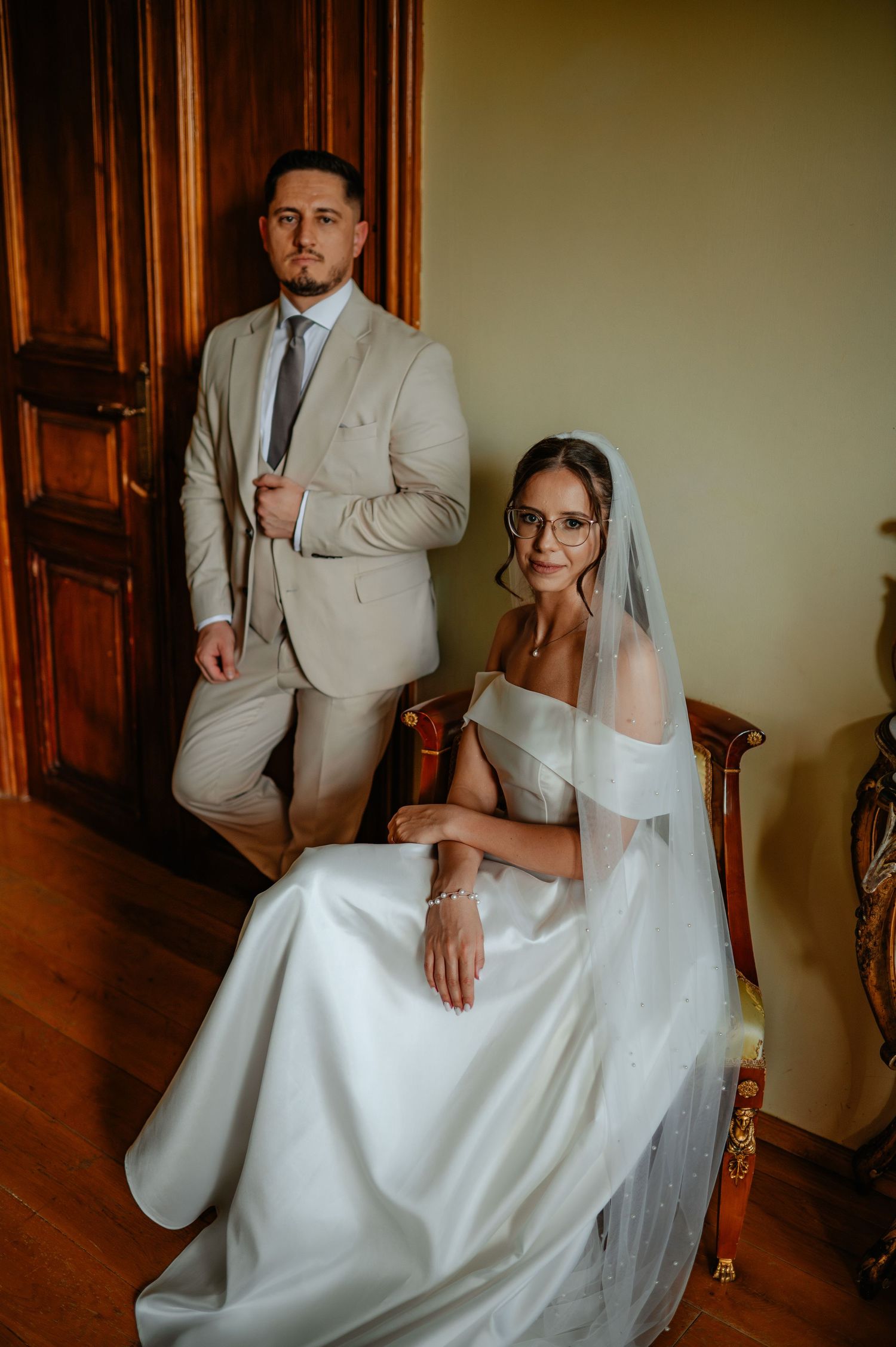 Bride in white off-shoulder gown with veil and groom in beige suit posing in elegant interior.