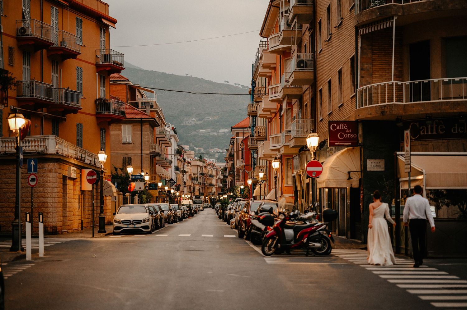 Couple in wedding attire strolling down an Italian street at dusk with lights.