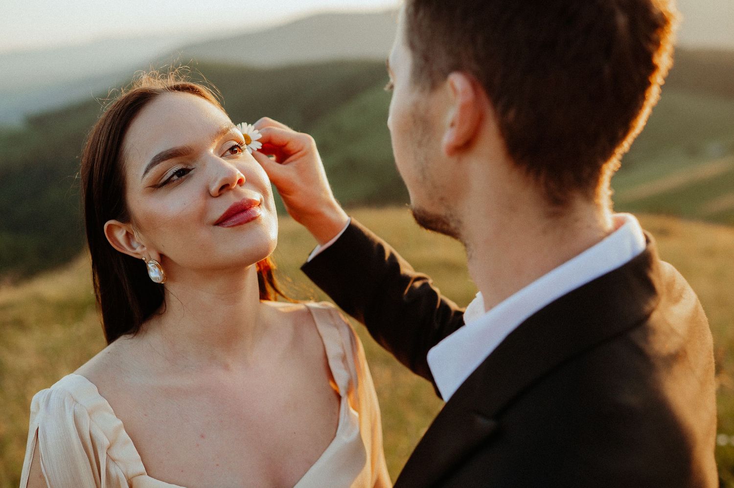 Groom tenderly touches brides face in a sunlit mountain field during romantic wedding photo session.