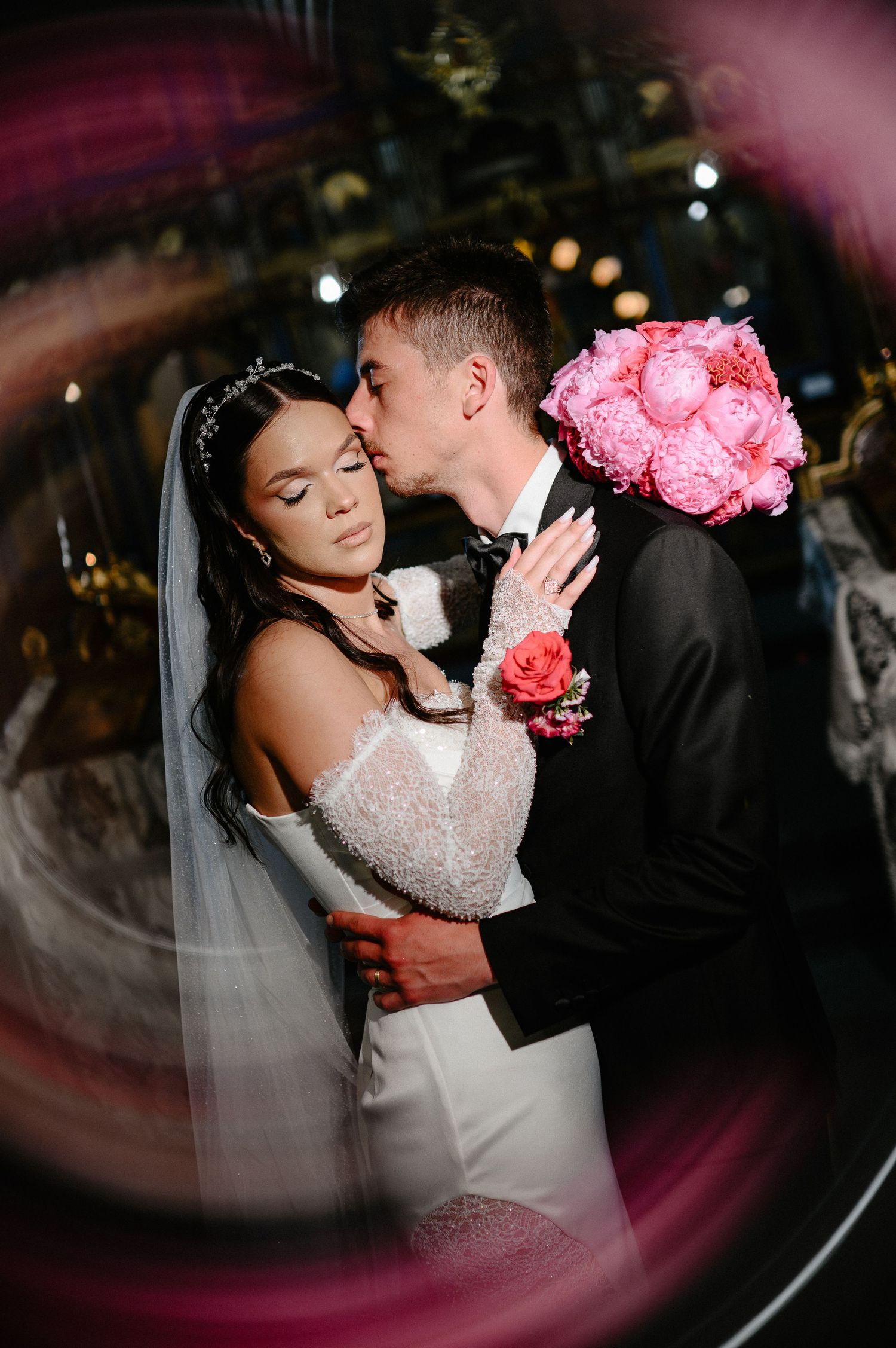 Artistic circular framed shot of groom kissing bride holding pink peony bouquet in Orthodox church.
