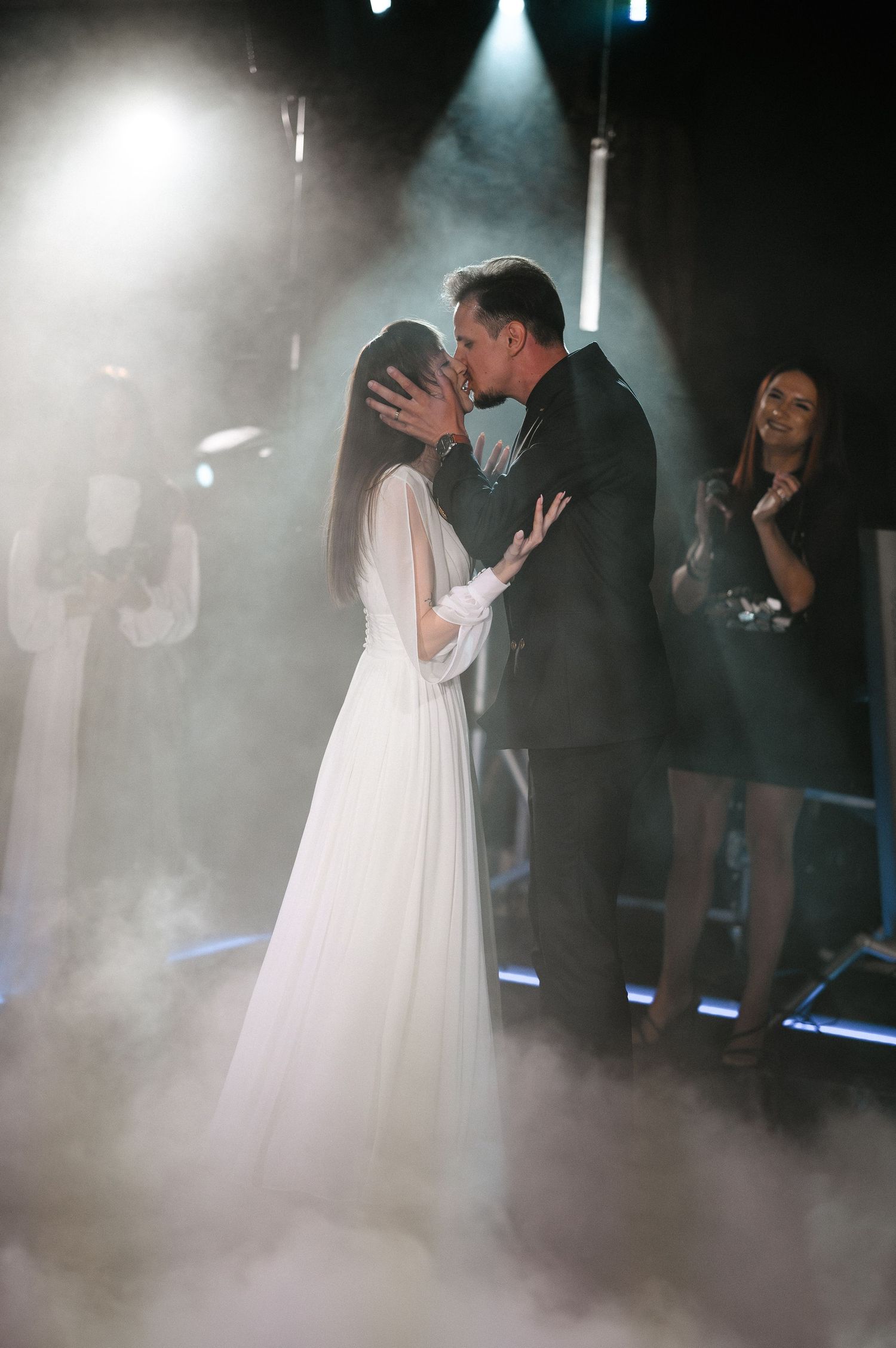 Bride and groom share first kiss on fog-covered dance floor under dramatic spotlights at reception.