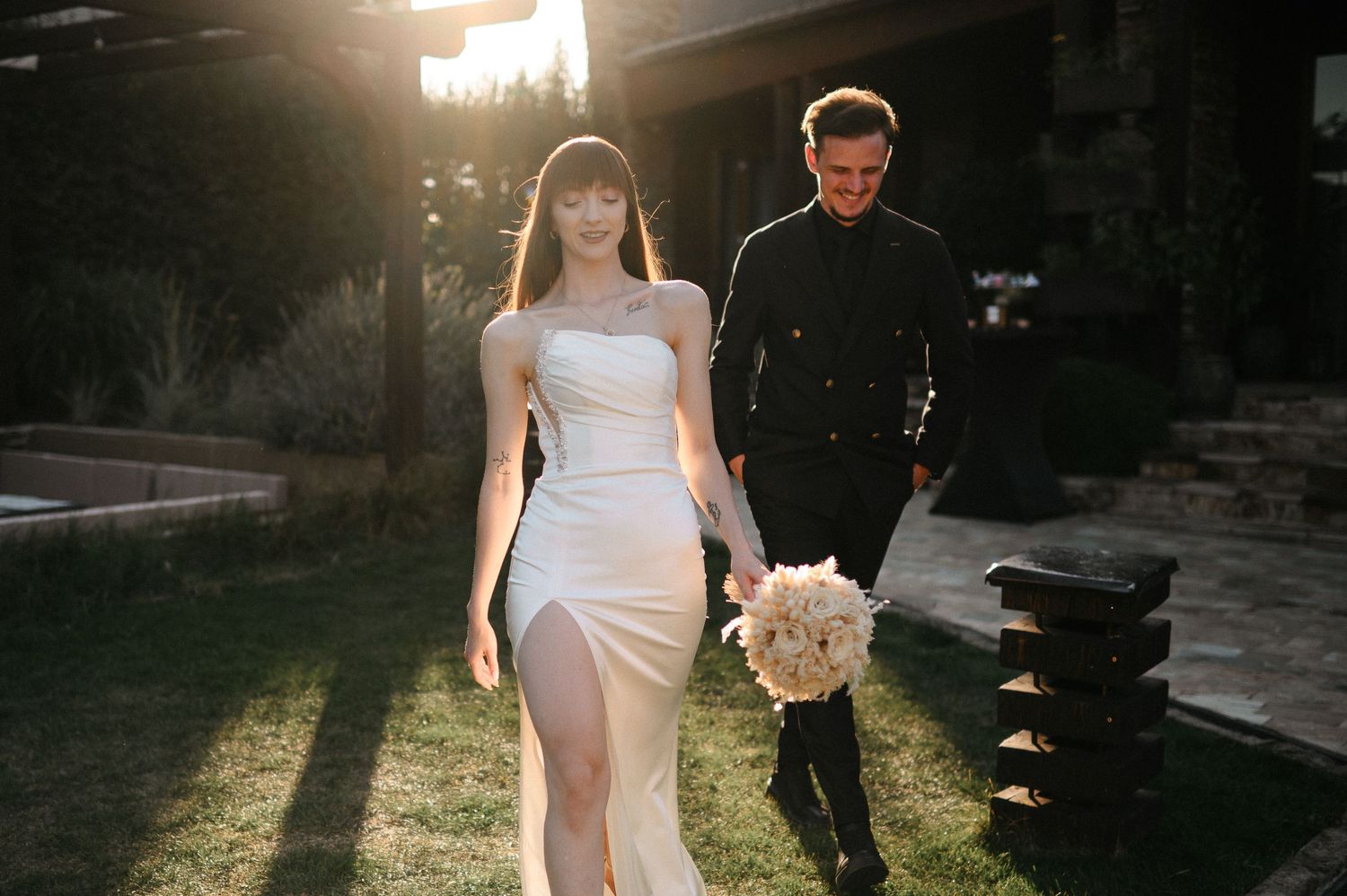 Bride in white slit gown holds bouquet walking with groom in black suit in golden sunset light.