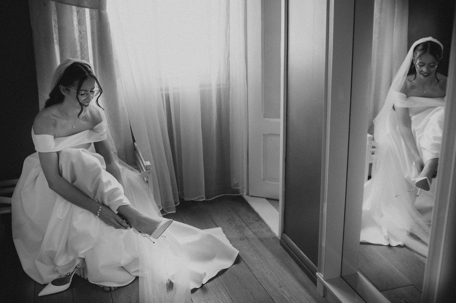 Bride in off-shoulder gown adjusting her shoe before mirror in black and white wedding preparation photo.