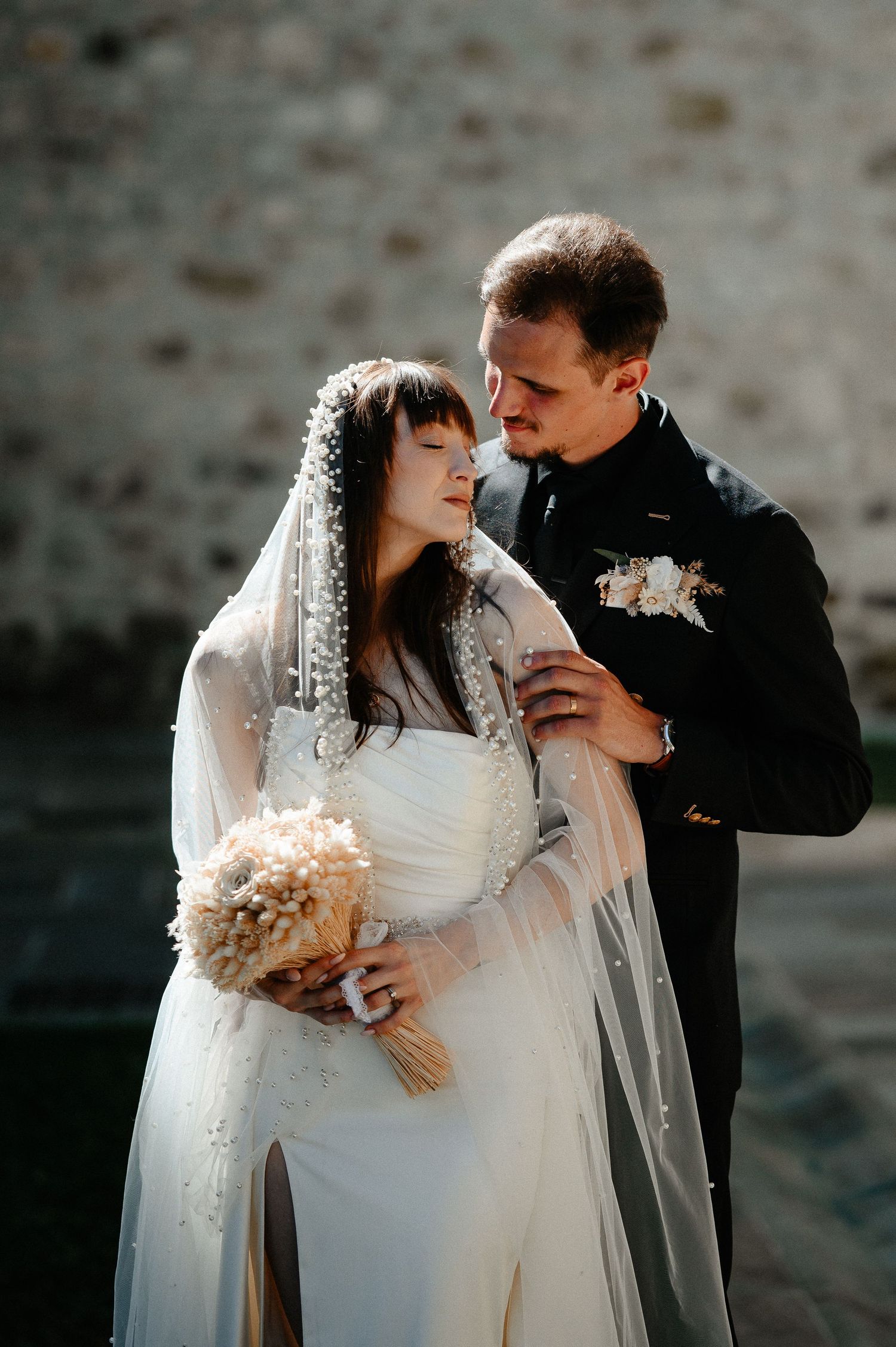 Bride in pearl veil and white gown embraces groom in black suit outdoors holding dried floral bouquet.