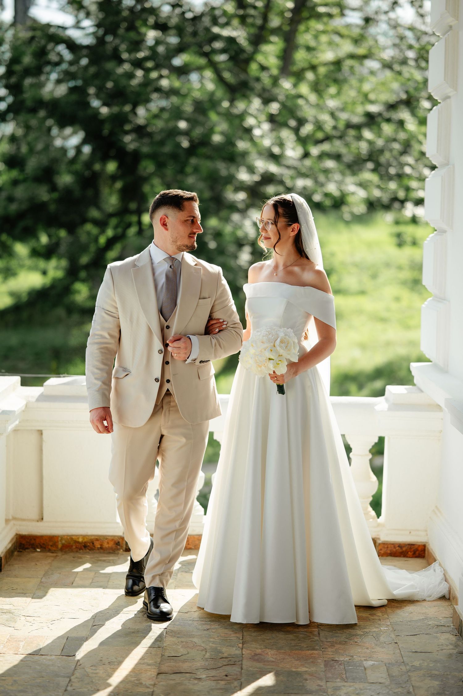 Bride in off-shoulder white gown and groom in beige suit walk together smiling on sunlit classical white balcony.
